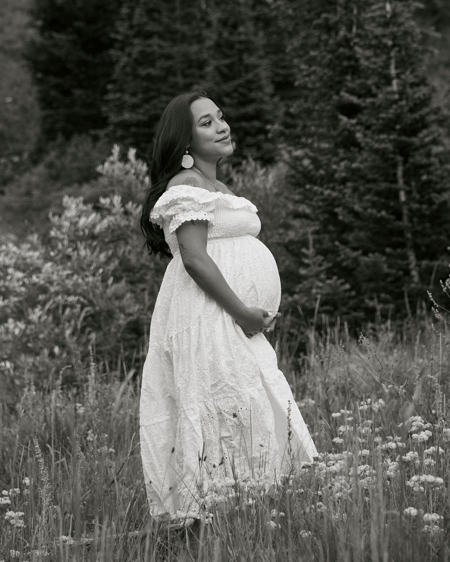 Black and white maternity portrait of person in lace dress standing in mountain wildflower field.