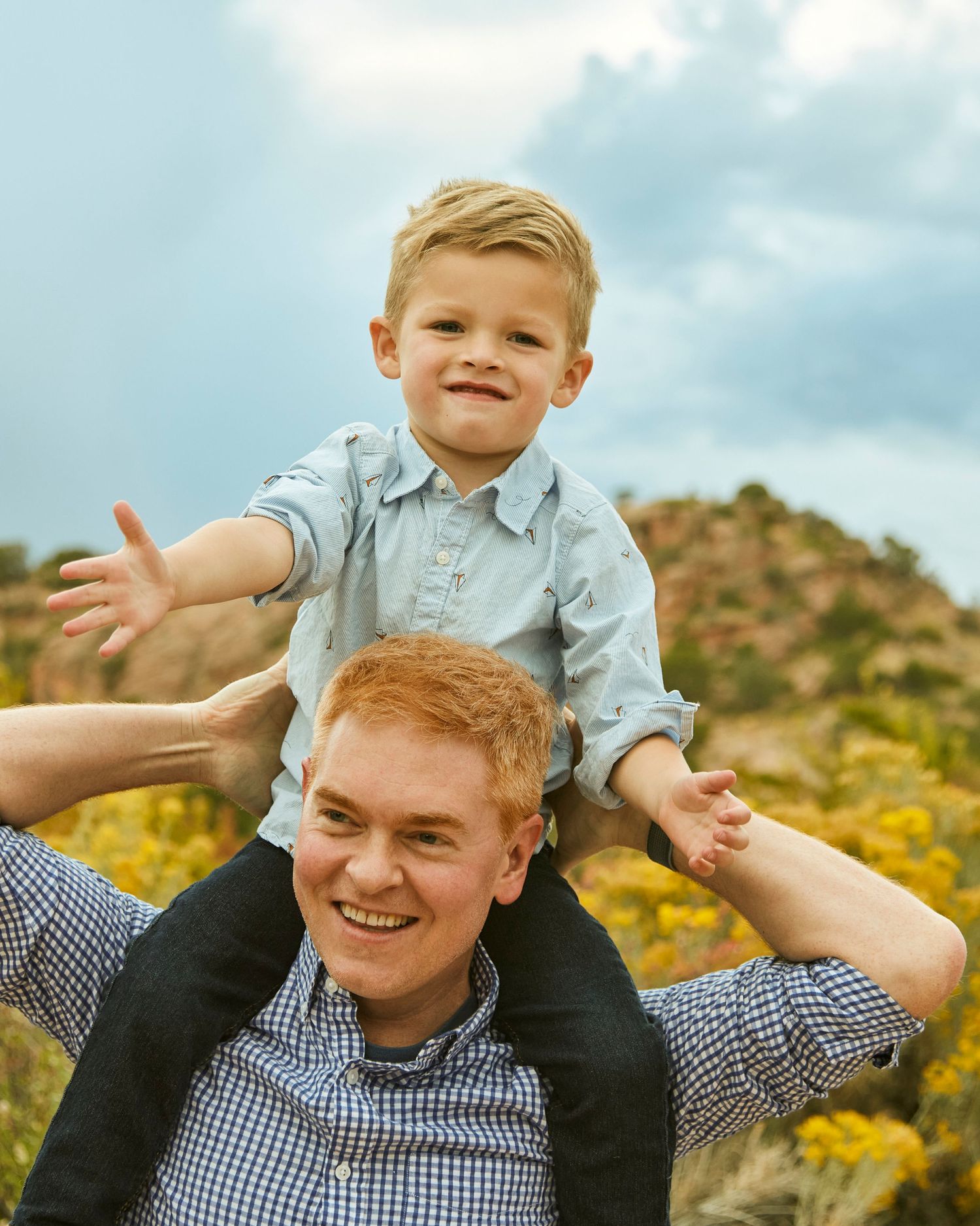 Father and child playing outdoors with yellow wildflowers and mountains in background.