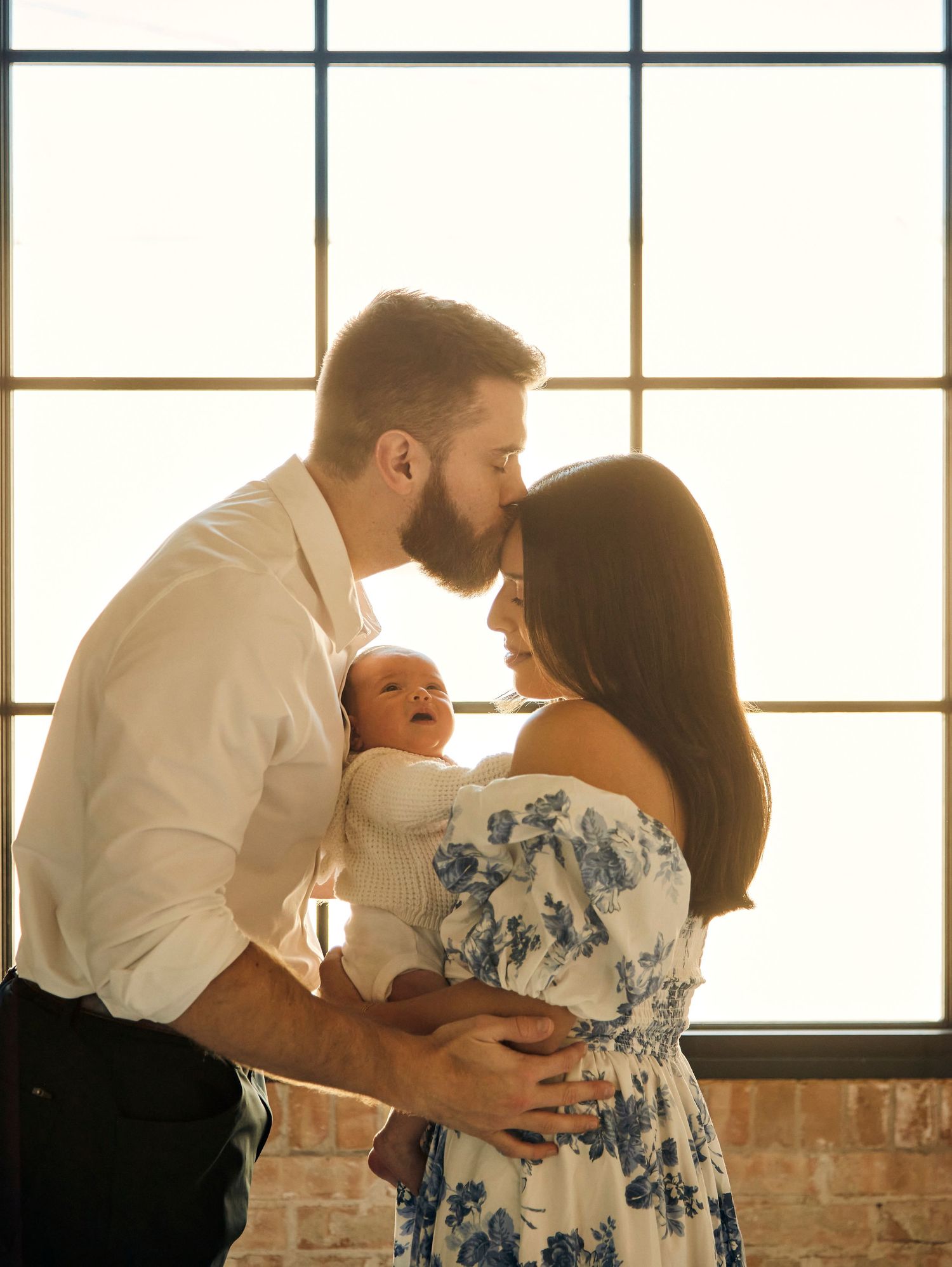 Tender family moment with newborn baby by sunlit window with brick wall.