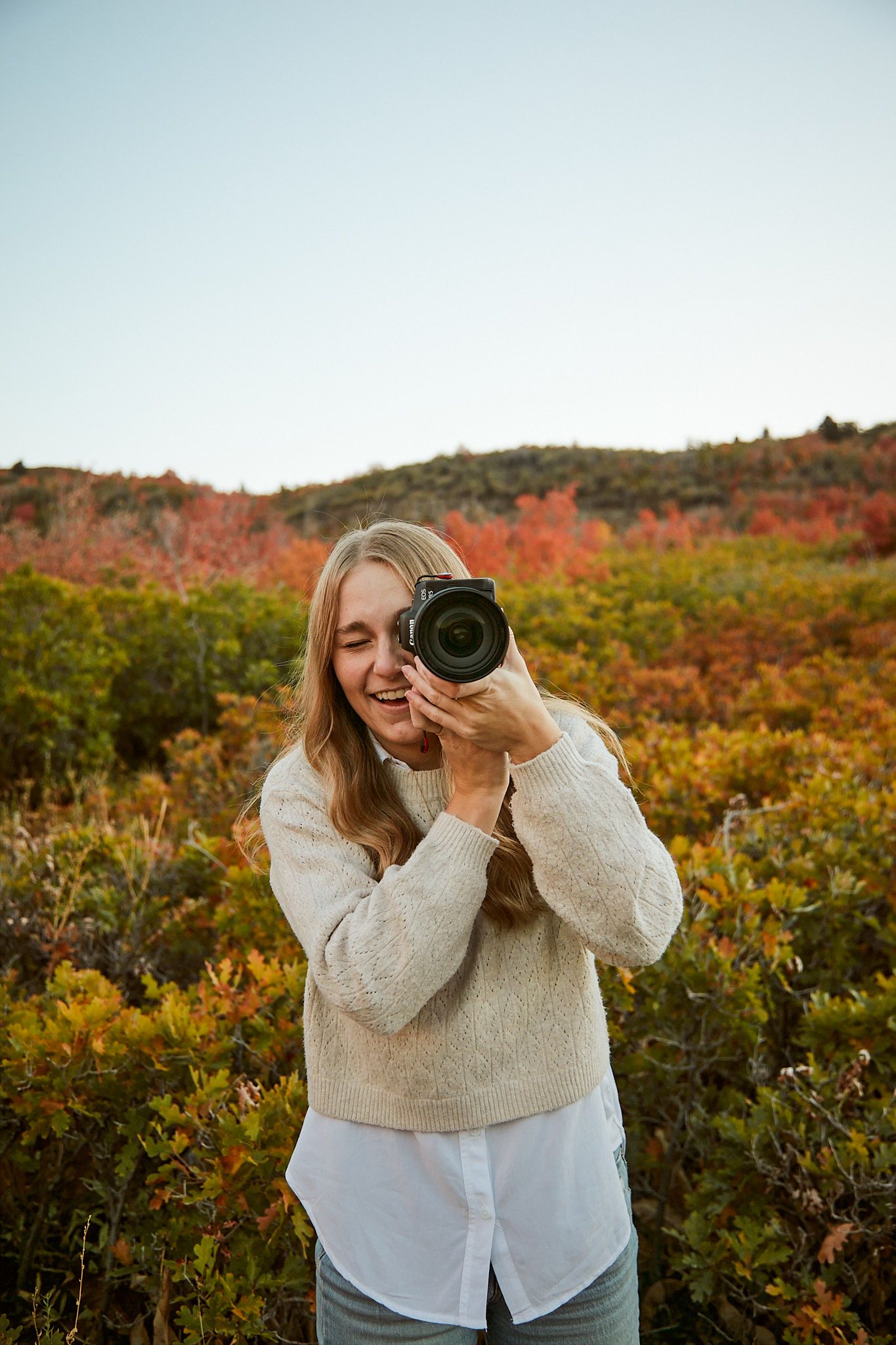 Photographer taking pictures among colorful fall bushes wearing layered cream and white outfit.