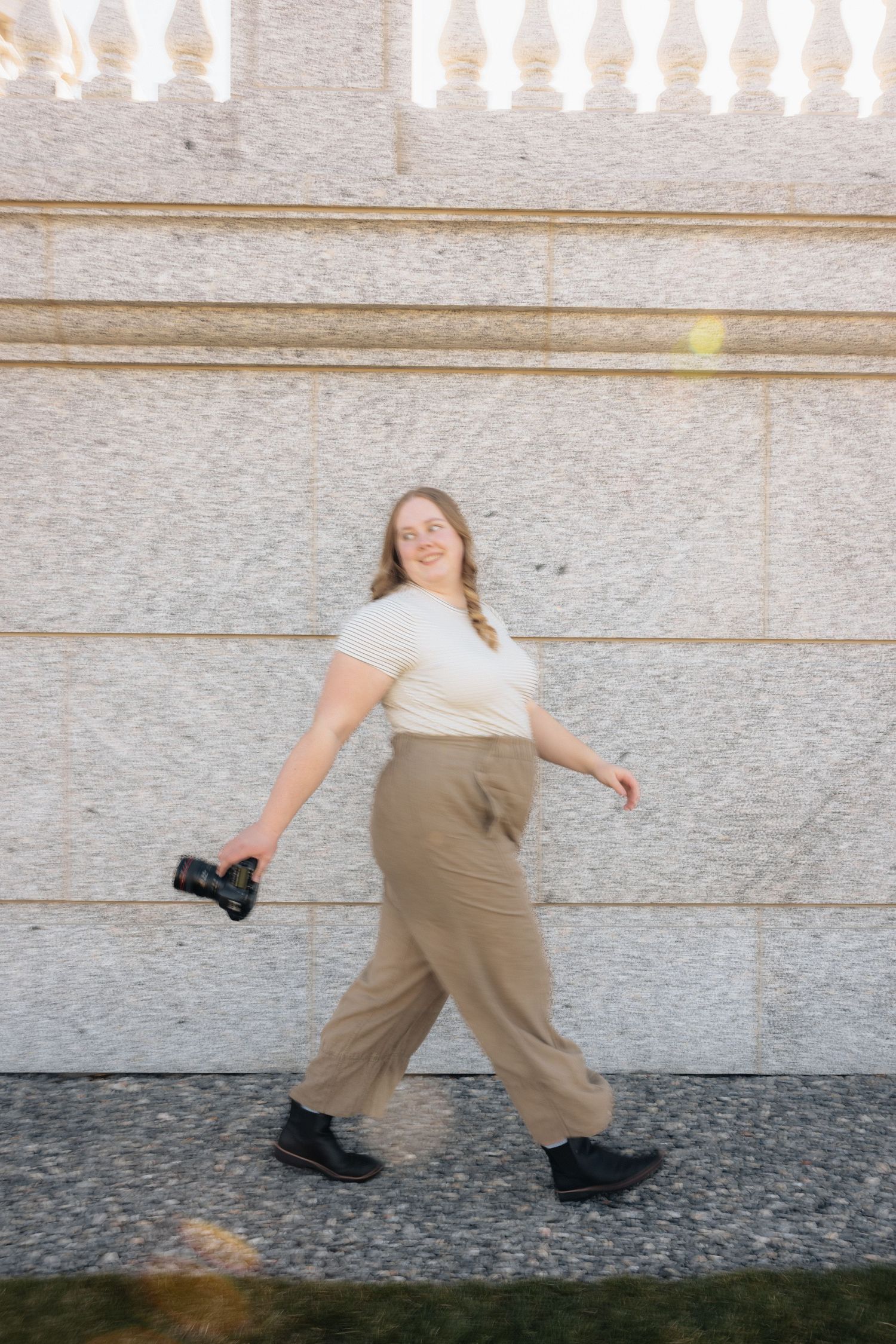 Person in white top and tan pants walking against stone wall while holding camera.