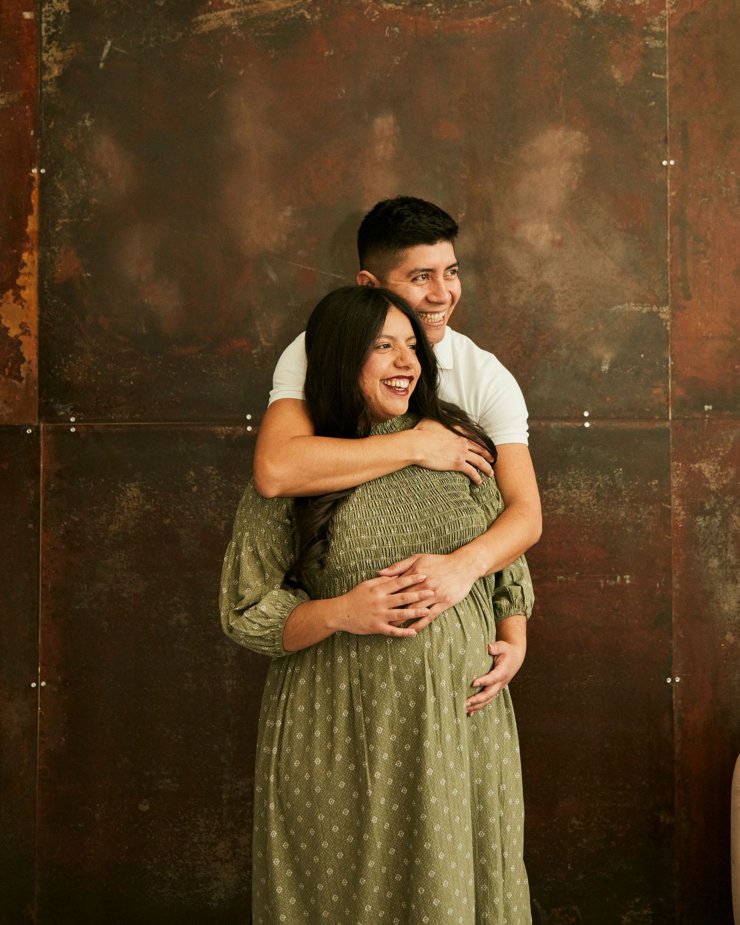 Couple embraces against rustic metal wall backdrop in casual and polka dot dress attire.