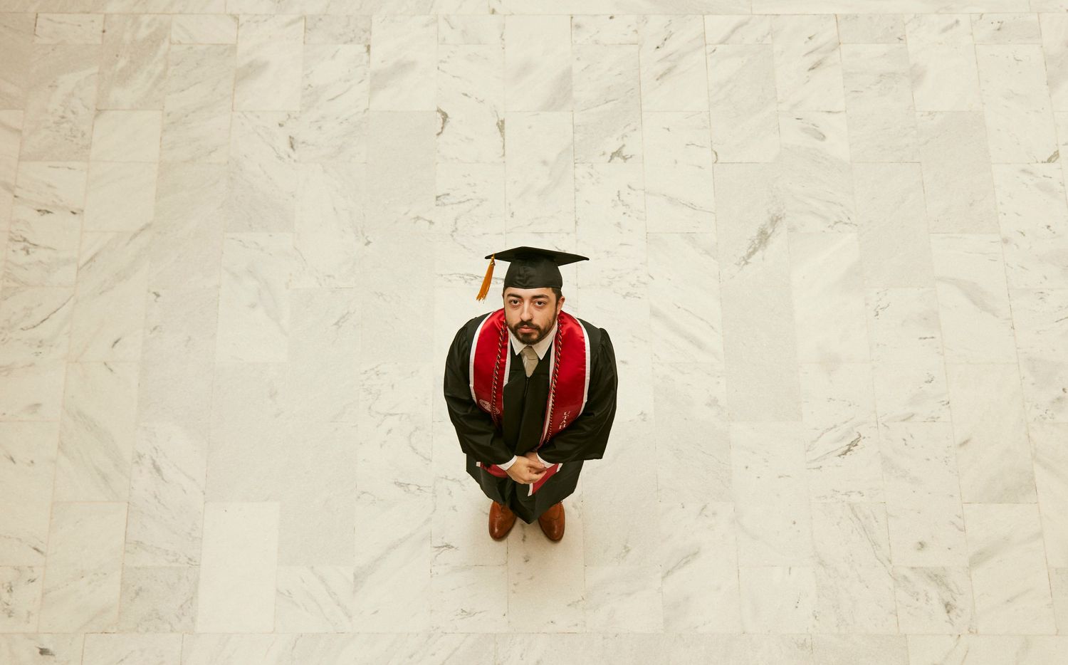 Graduate in cap and gown poses from above on white marble floor.