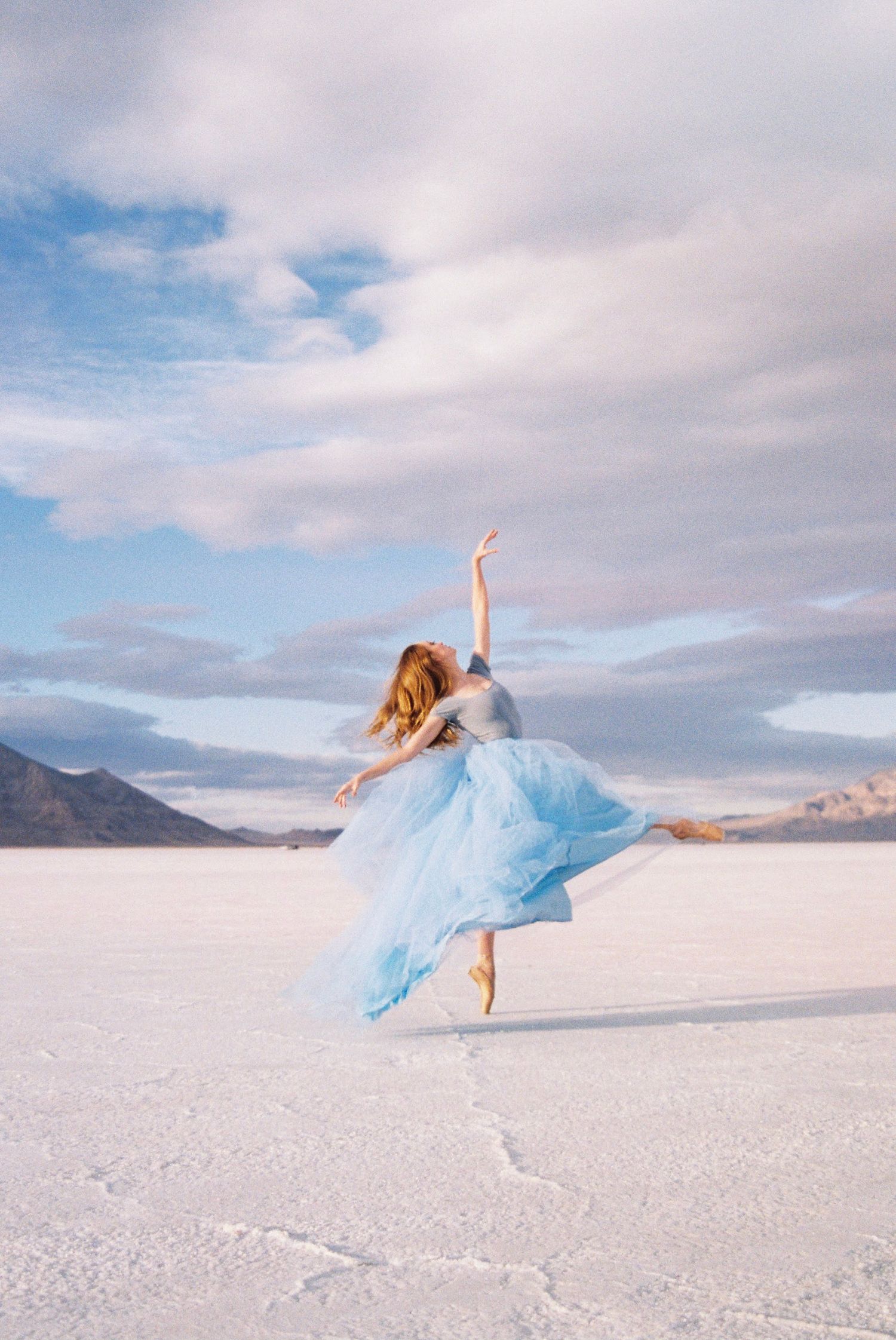 A dancer in a flowing blue dress performs on the salt flats against a cloudy mountain backdrop.
