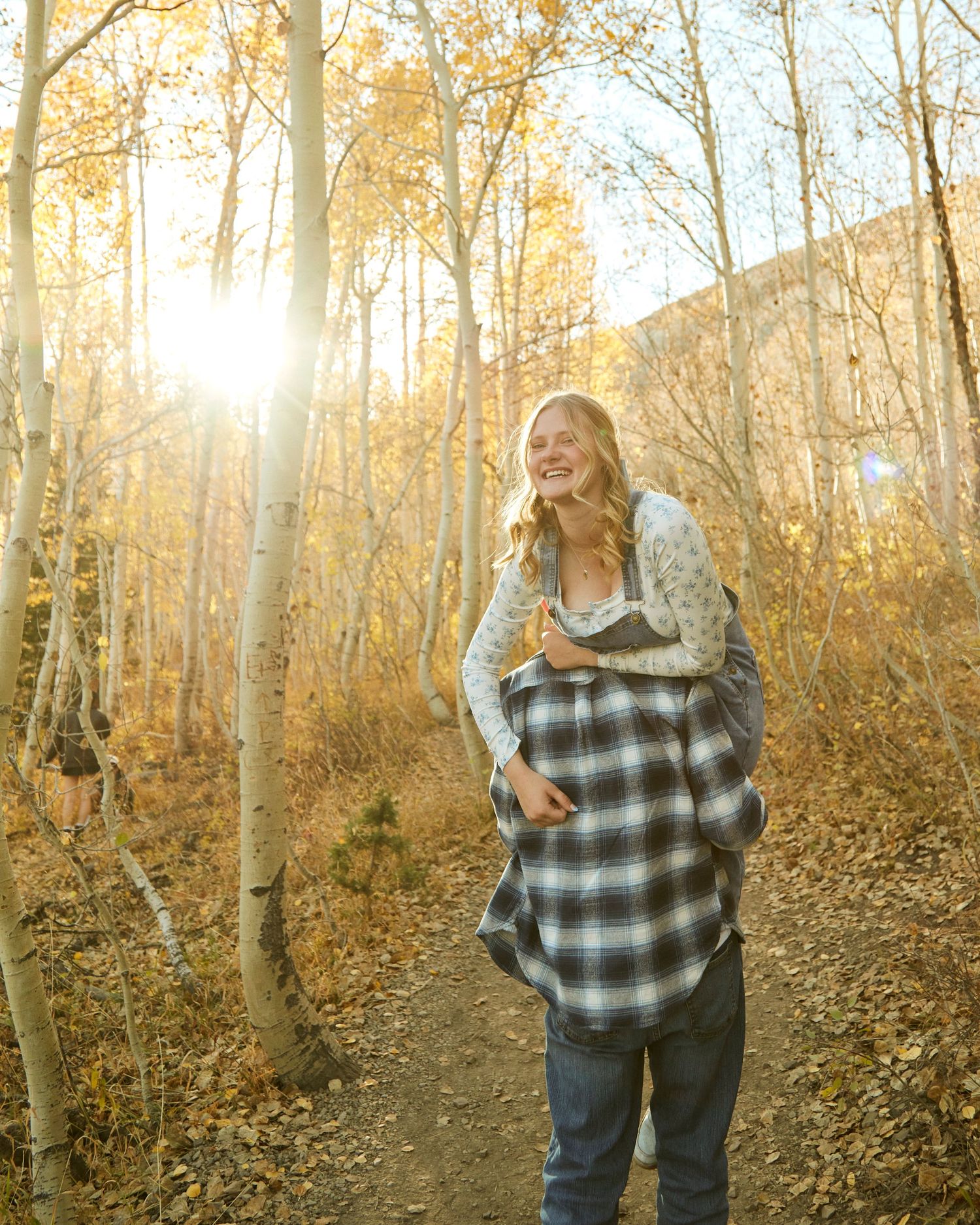 Person displaying engagement ring while laughing in a sun-lit autumn forest.