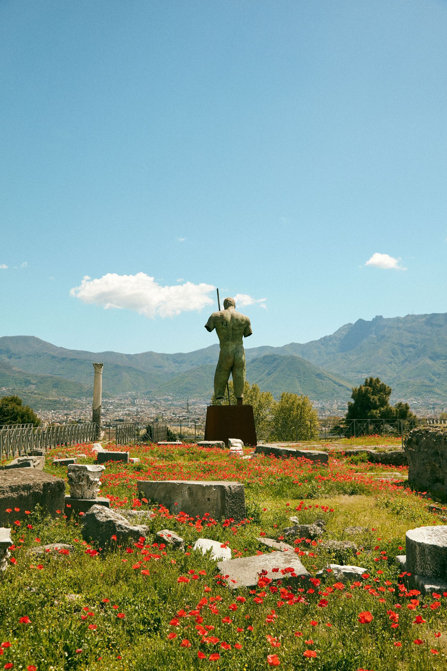 Bronze statue stands among red wildflowers with mountains in background under blue sky.