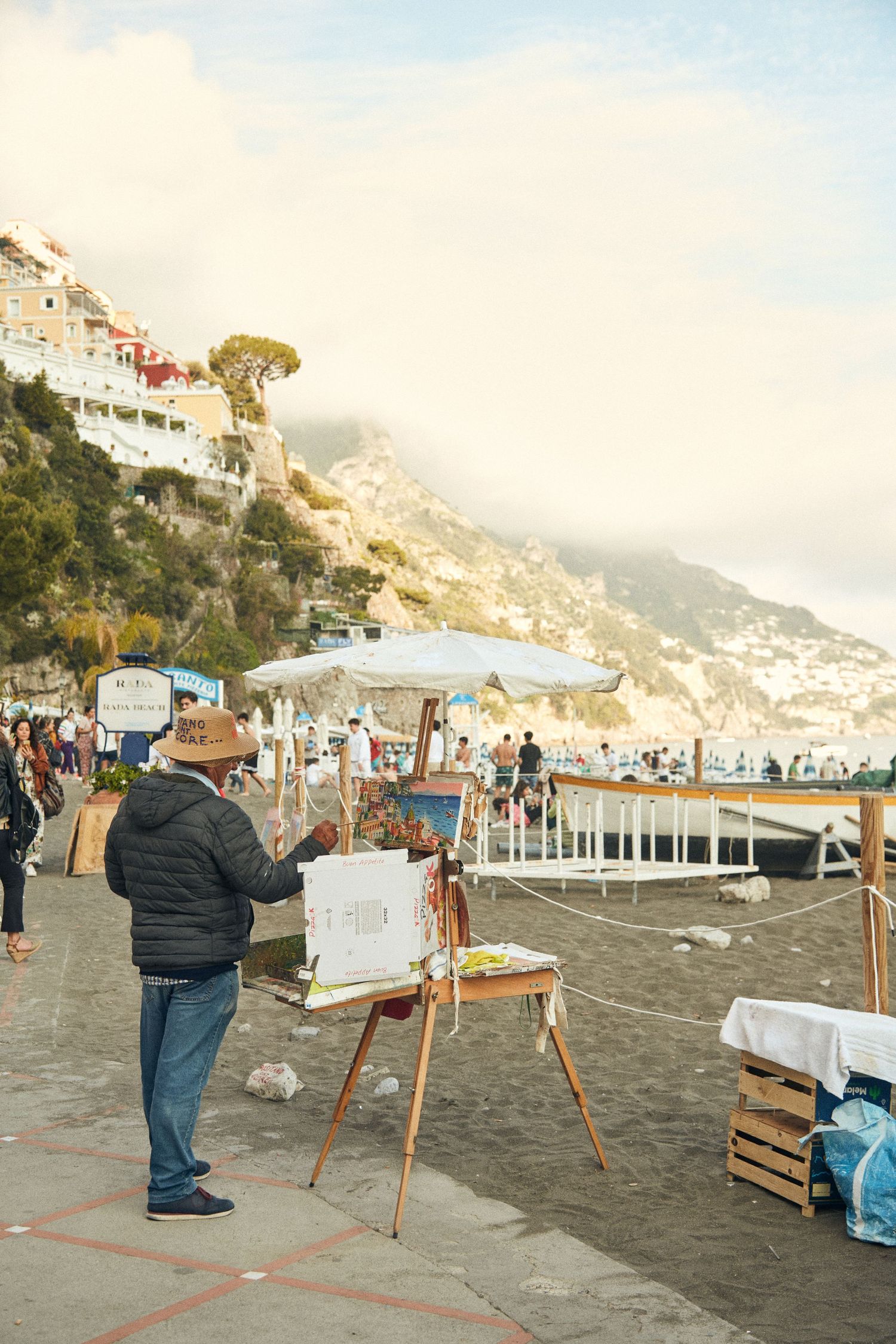 Artist painting on easel at beach promenade with Positano's hillside architecture in background.