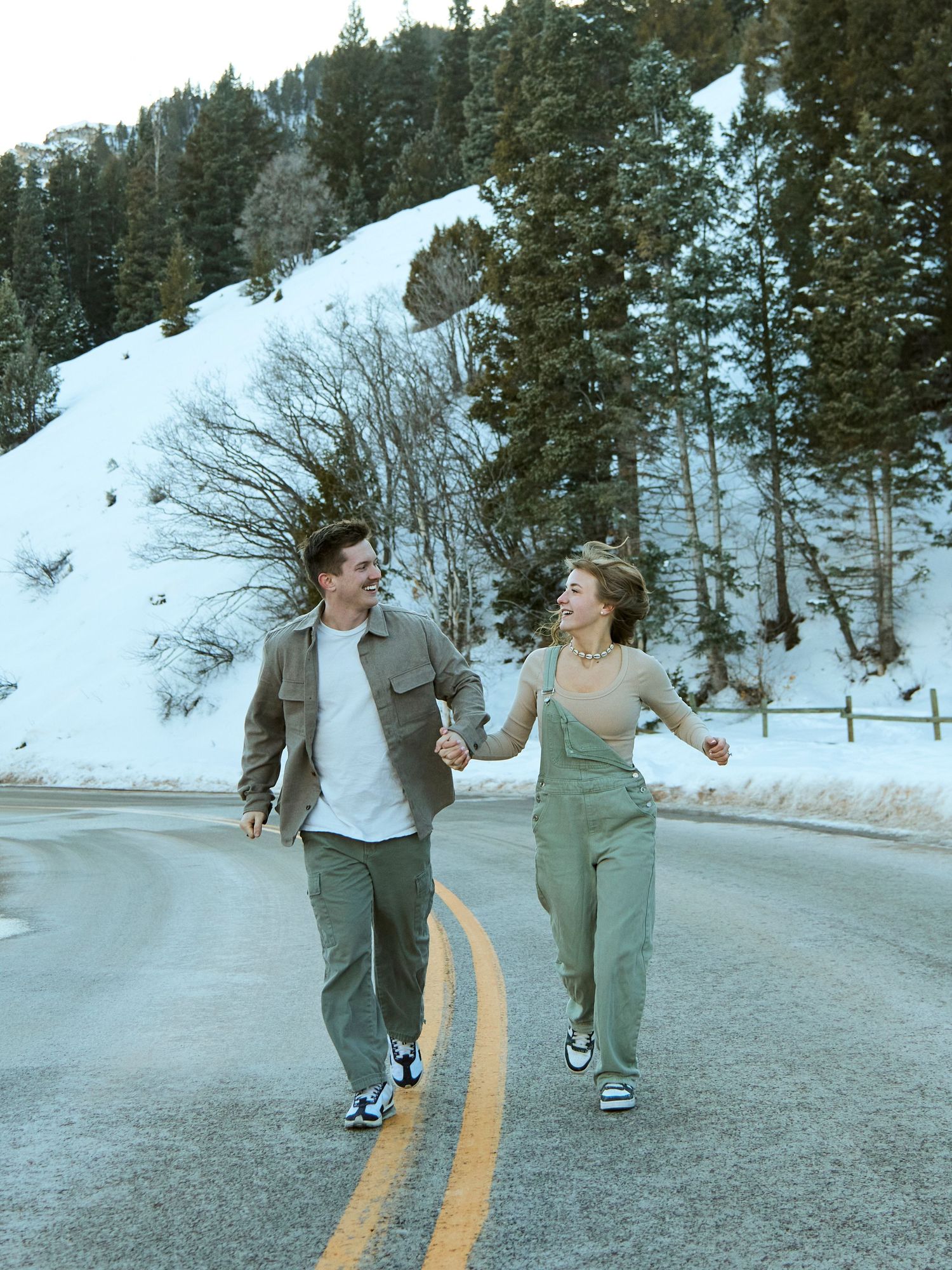 Happy couple running on a mountain road surrounded by snow and pine trees.