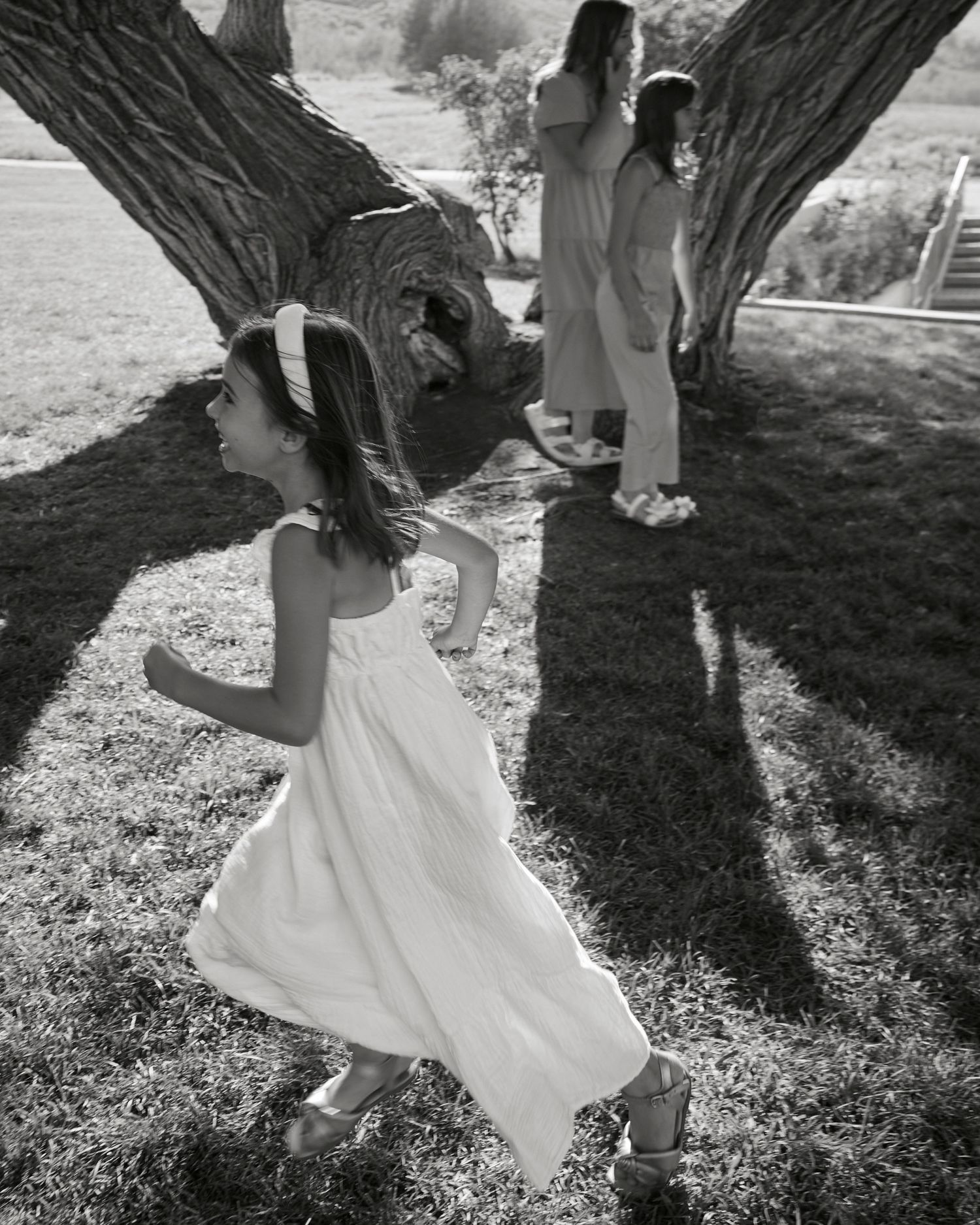 Black and white photo of a child dancing and twirling in a white dress under a large tree.
