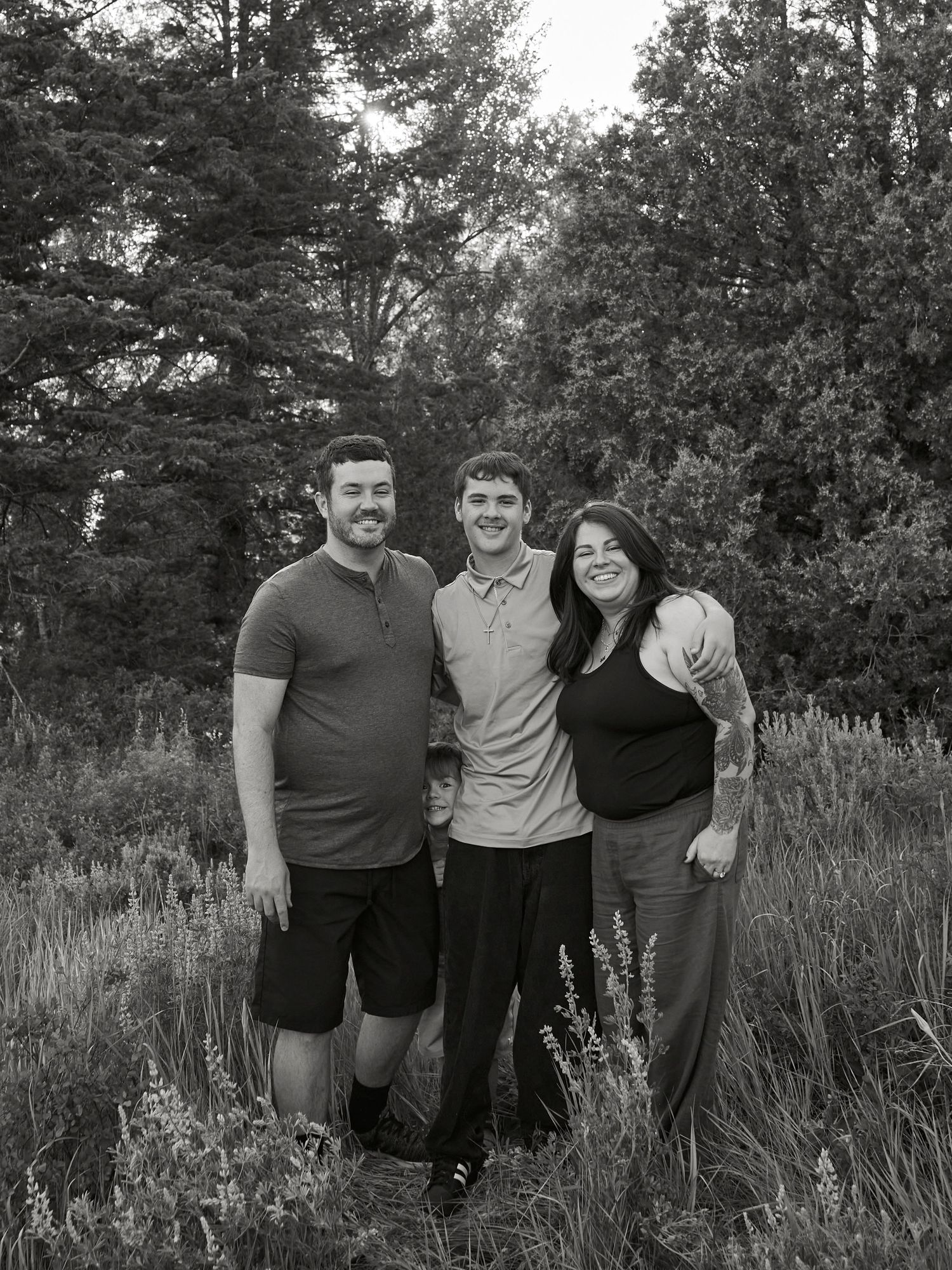 Family portrait in black and white with three people standing together in a meadow with trees in background.