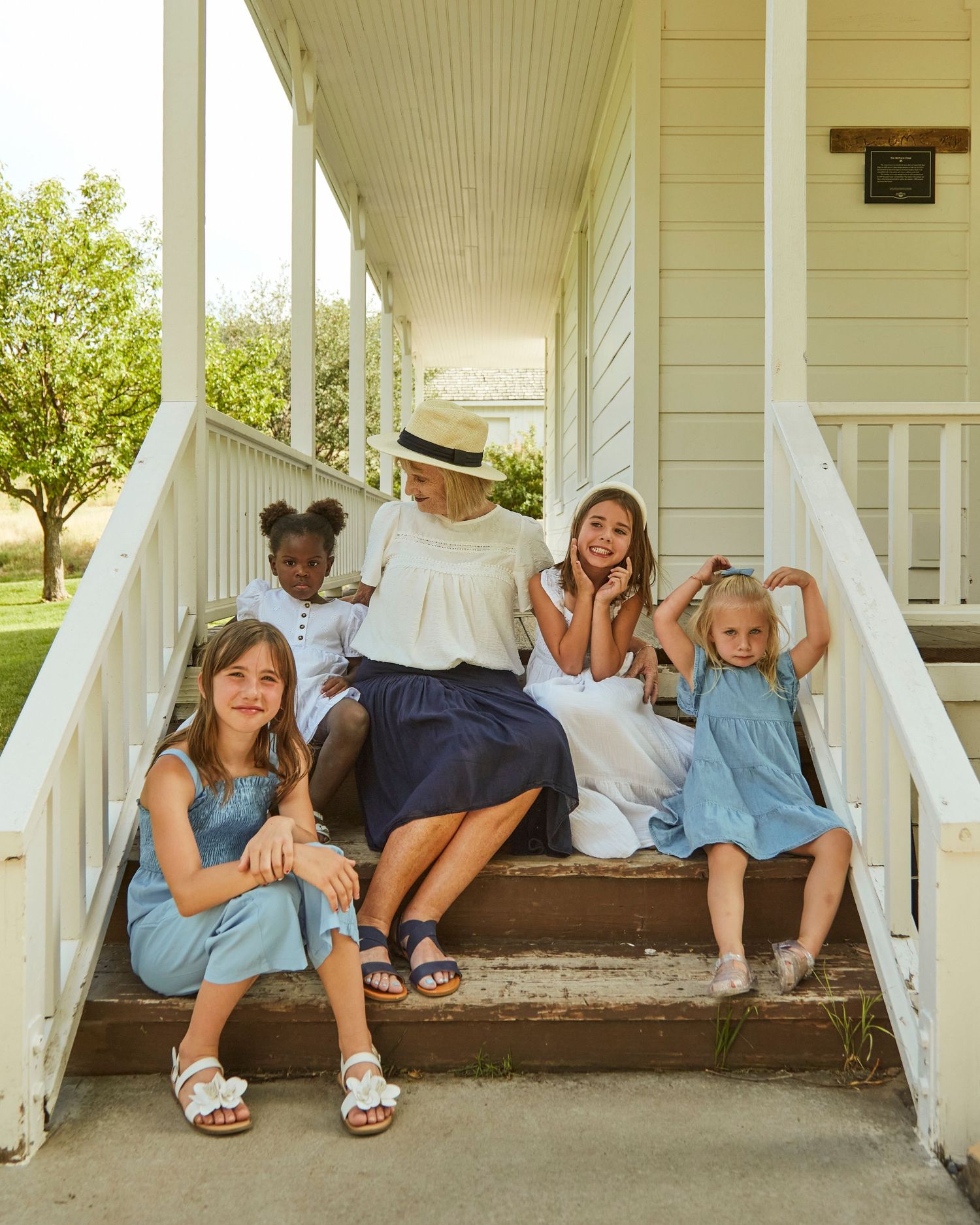 A cheerful group poses together on white farmhouse porch steps on a sunny summer day.