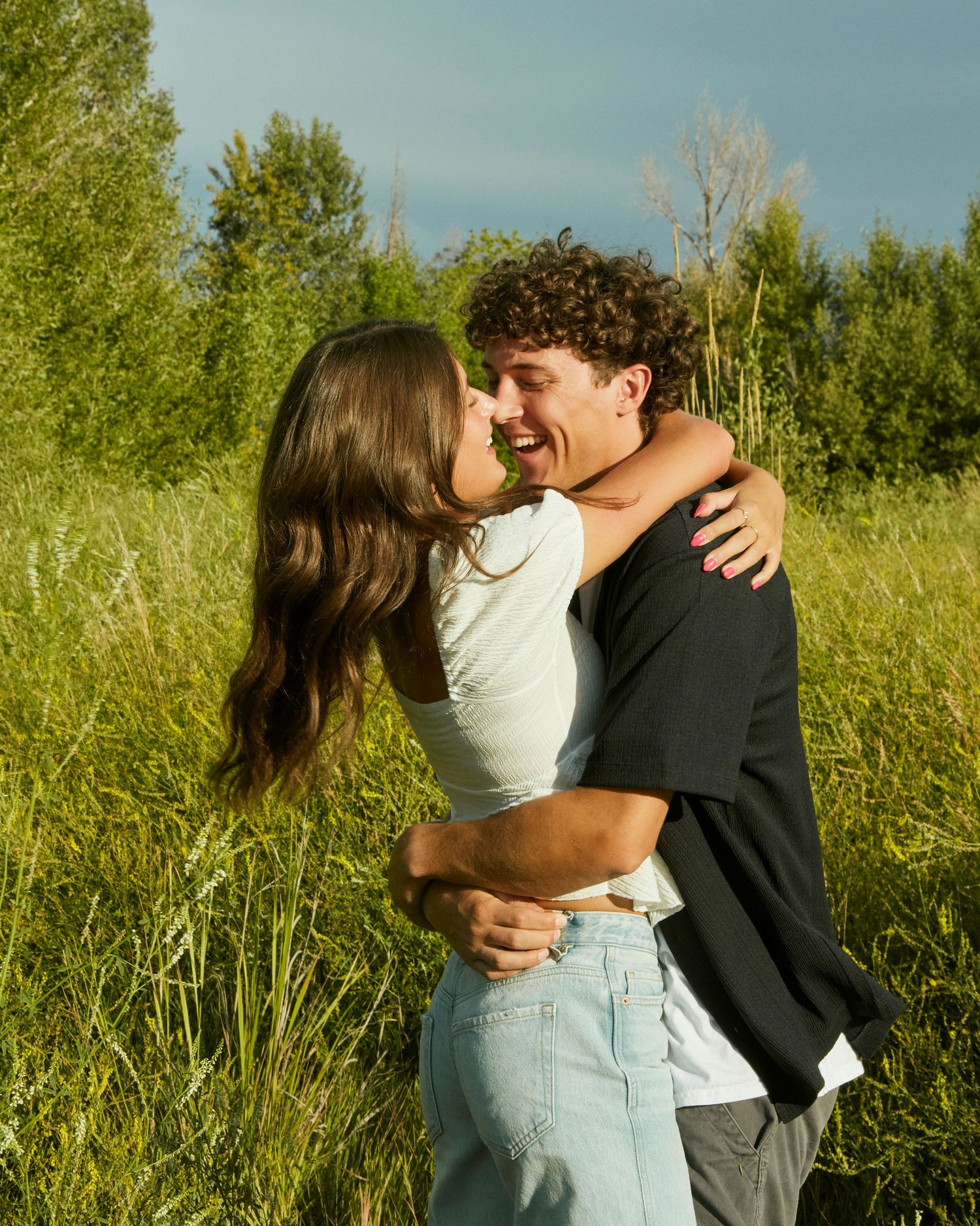 A couple embraces tenderly in a sunlit meadow with tall grass and trees in the background.