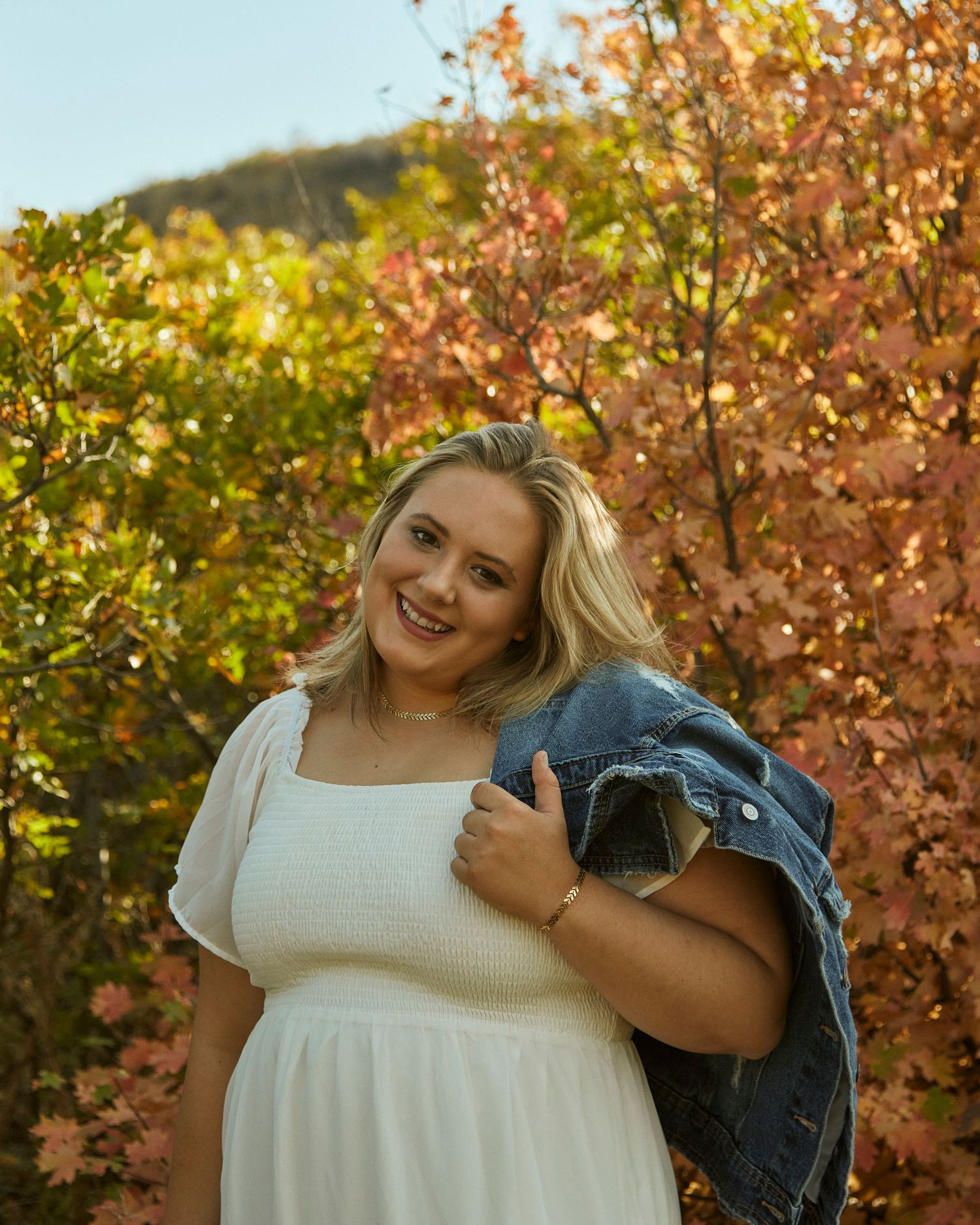 Person in white dress holding denim jacket against autumn foliage.