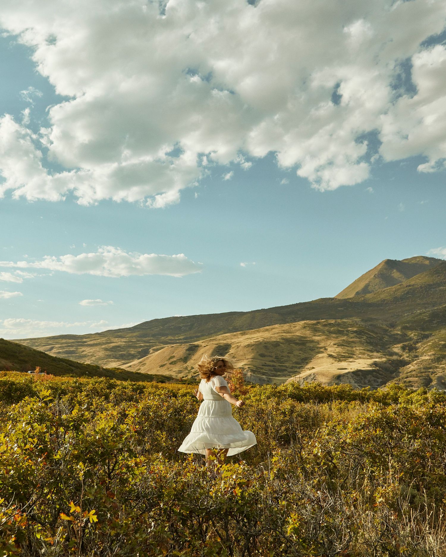 Figure in white dress twirling in mountain meadow under cloudy blue sky.
