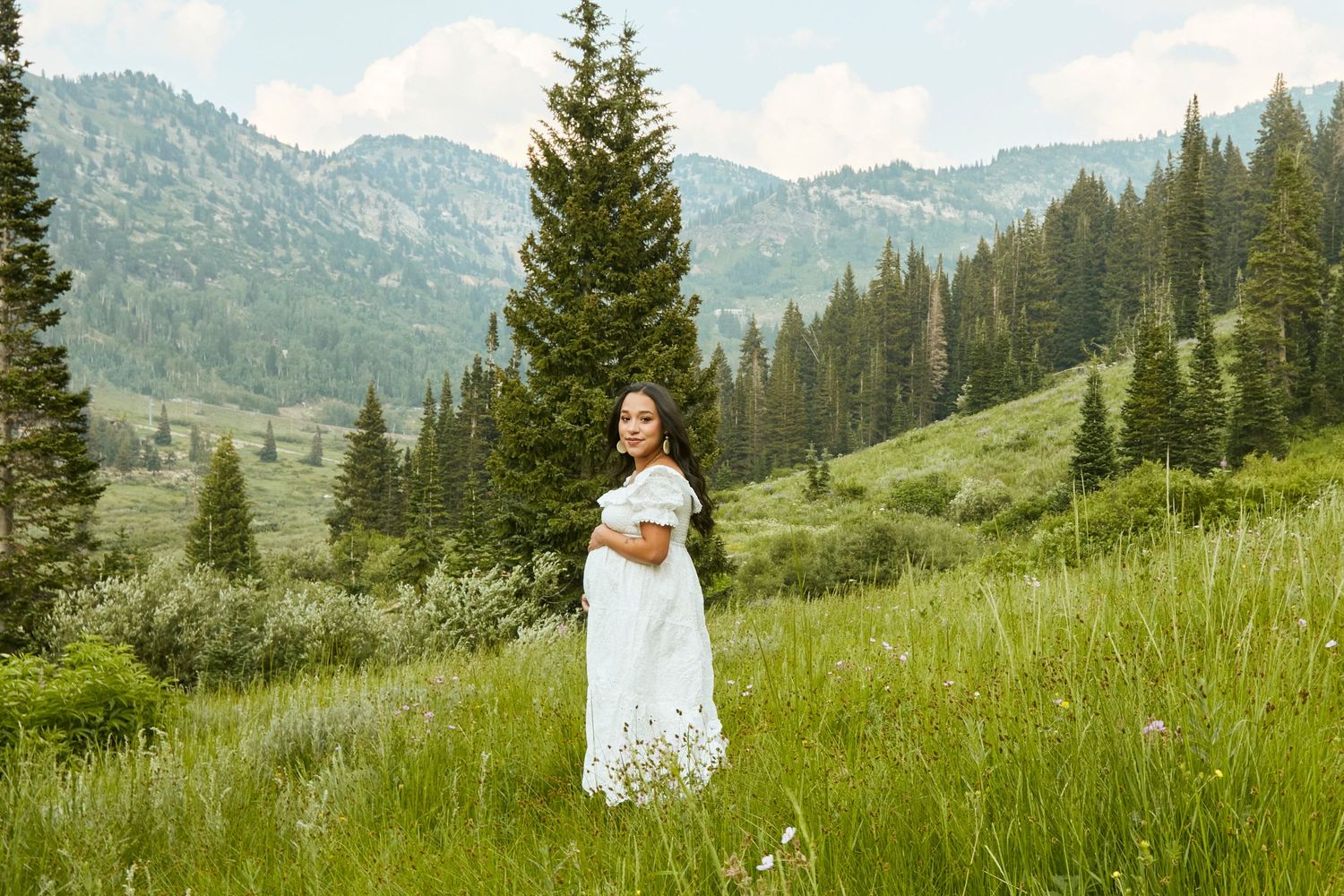A dreamy mountain maternity portrait with evergreen trees and distant peaks in the background.