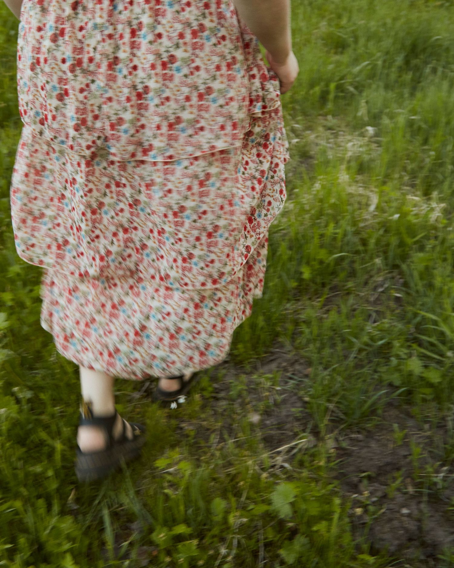 Close-up of floral dress hem and sandals walking on green grass path.
