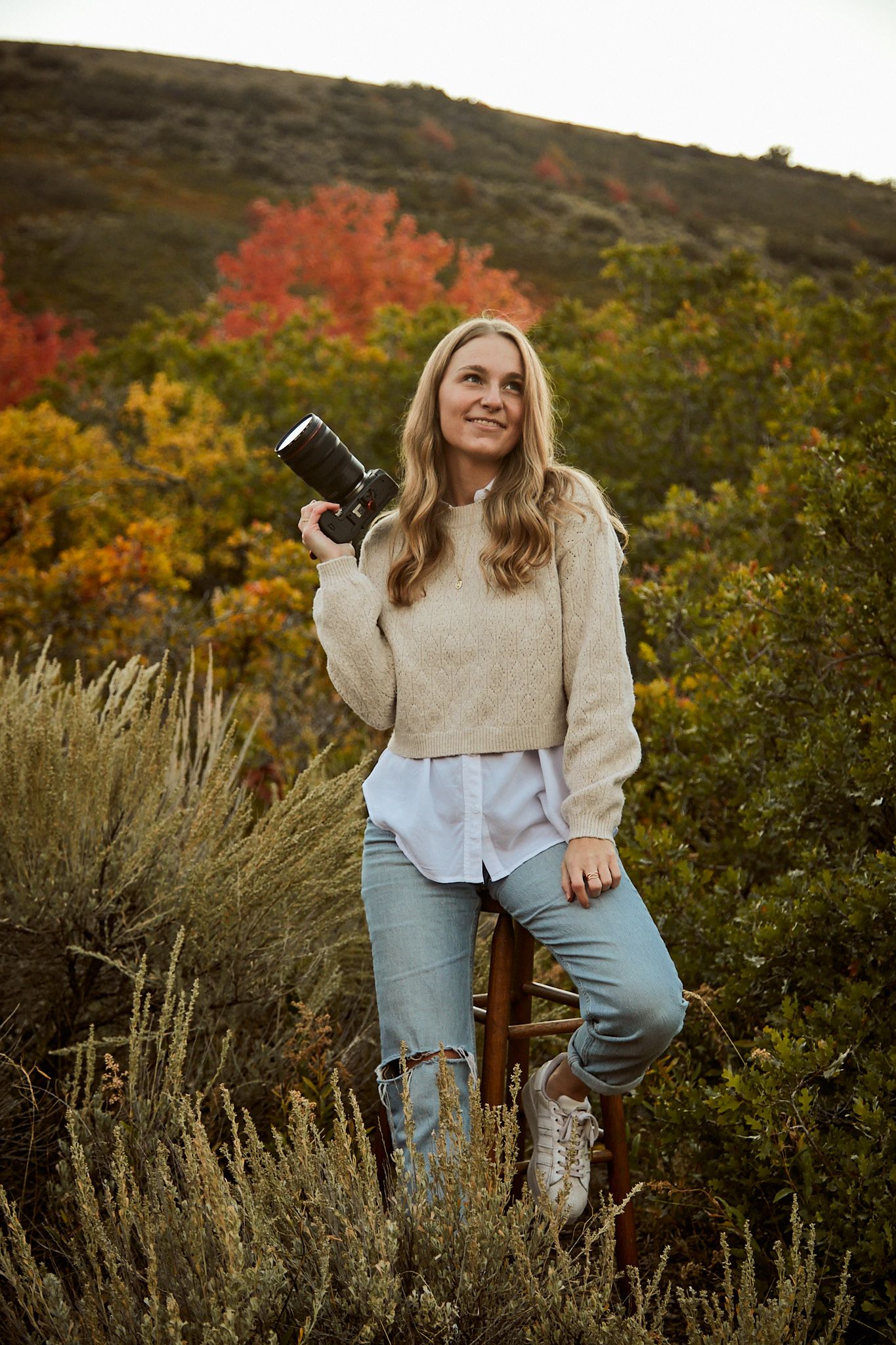 Photographer in casual autumn attire holds a camera while standing among colorful fall foliage and brush.