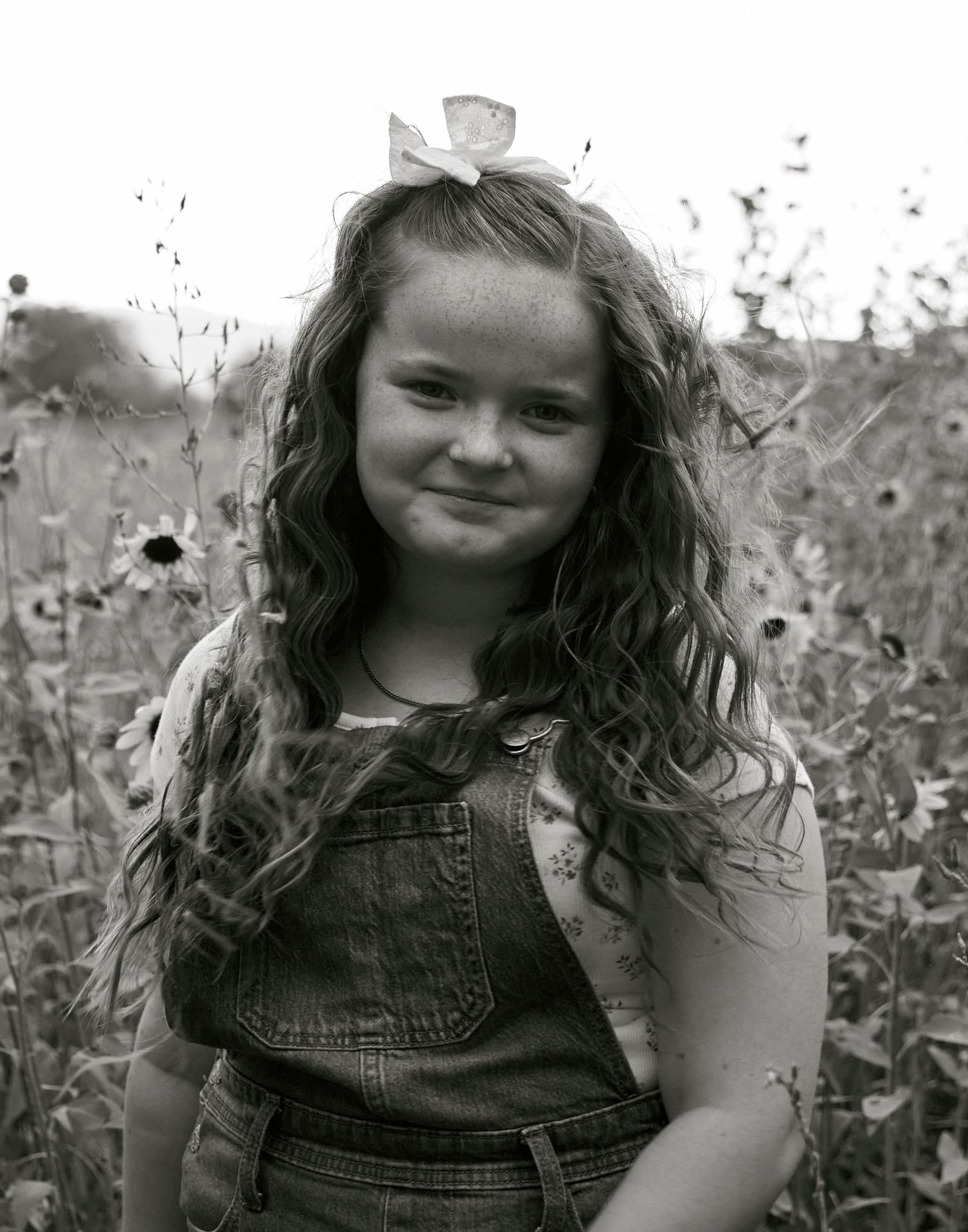 Black and white portrait of a young person with wavy hair wearing denim overalls in a wildflower field.