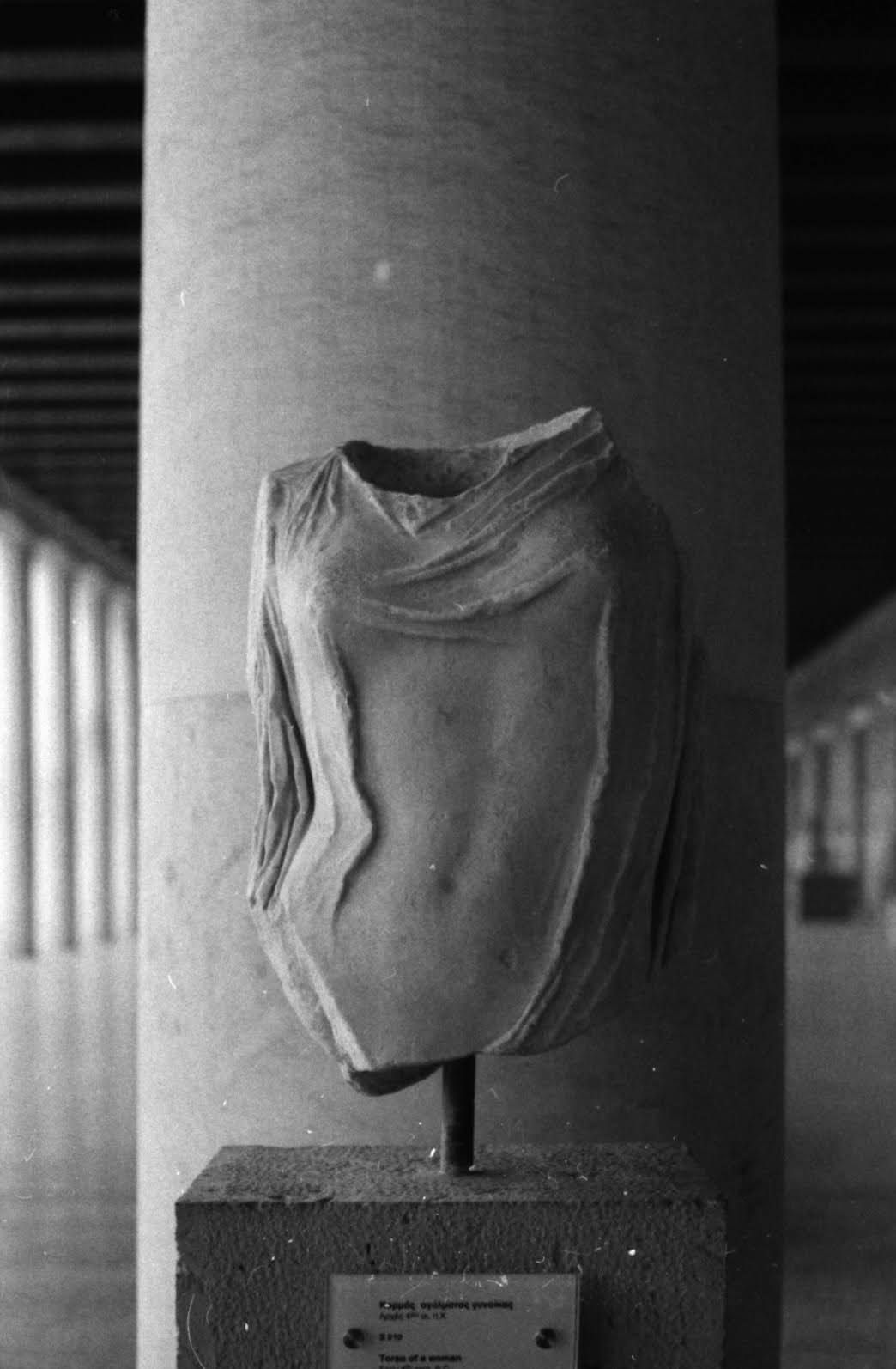 Black and white photograph of a draped torso sculpture displayed on pedestal in gallery.