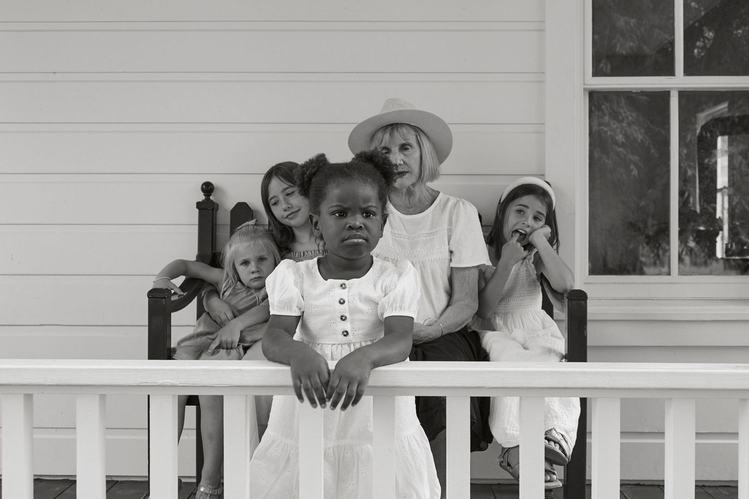 A vintage-style black and white portrait of a group sitting on a white porch railing.