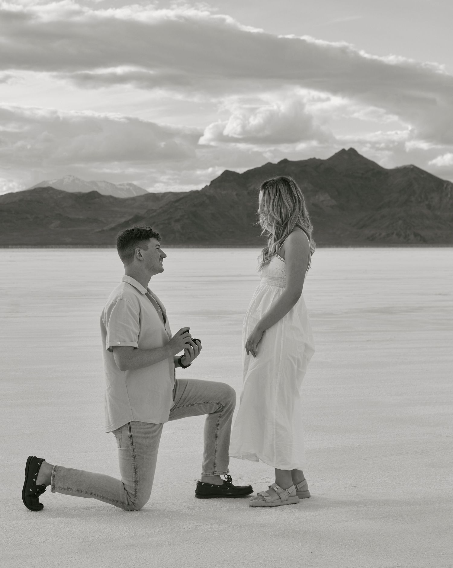 A black and white photograph of a romantic proposal moment on salt flats with mountains behind.