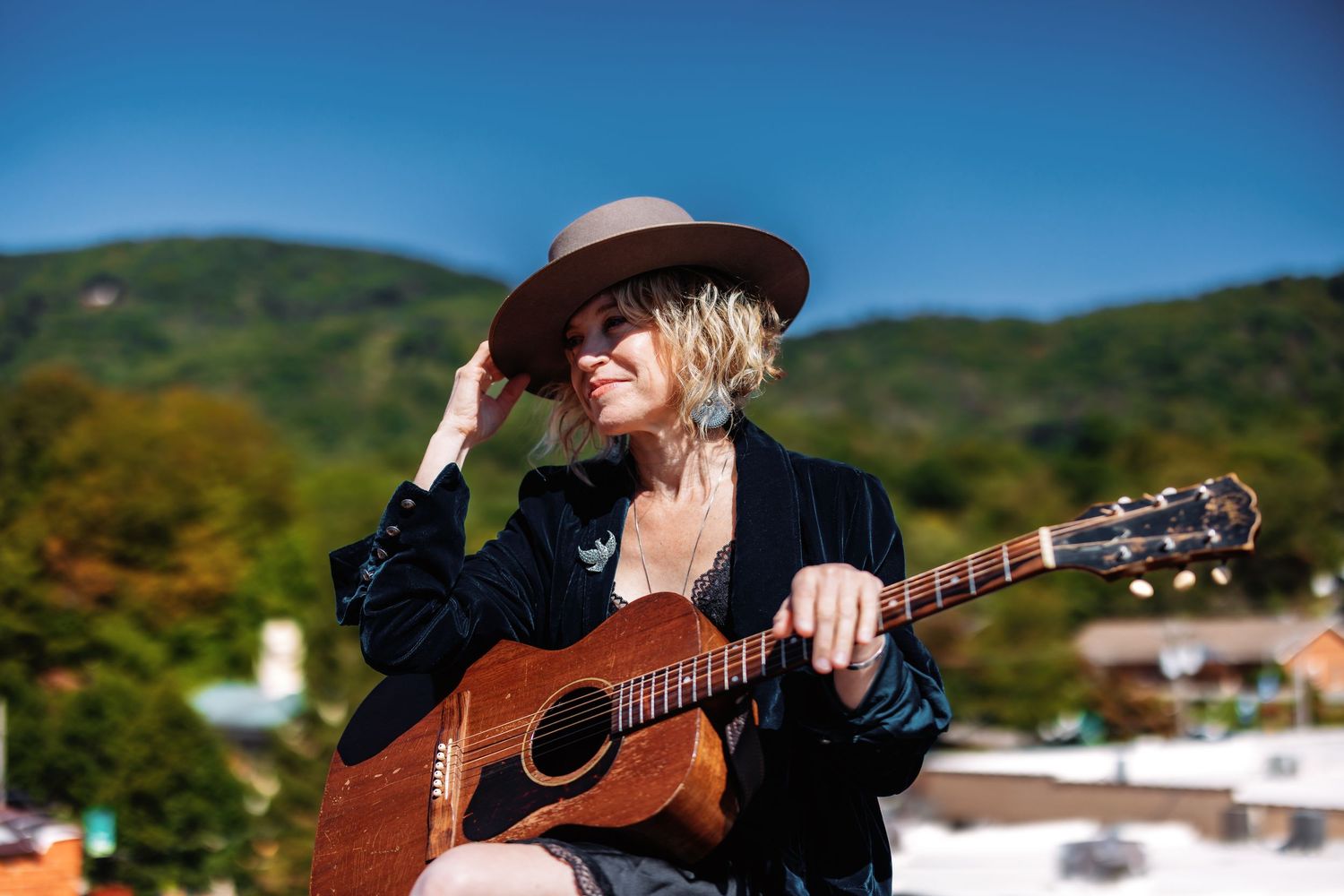 A musician plays an acoustic guitar outdoors against a scenic mountain backdrop on a sunny day.