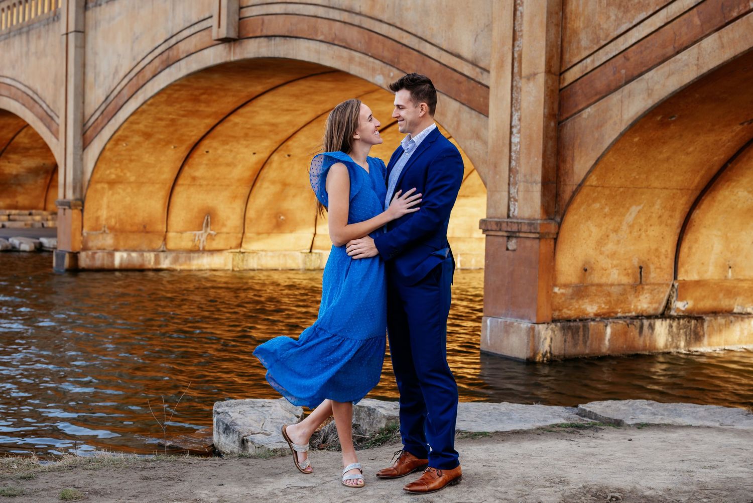 A couple in coordinating blue outfits embraces near an ornate bridge over water.