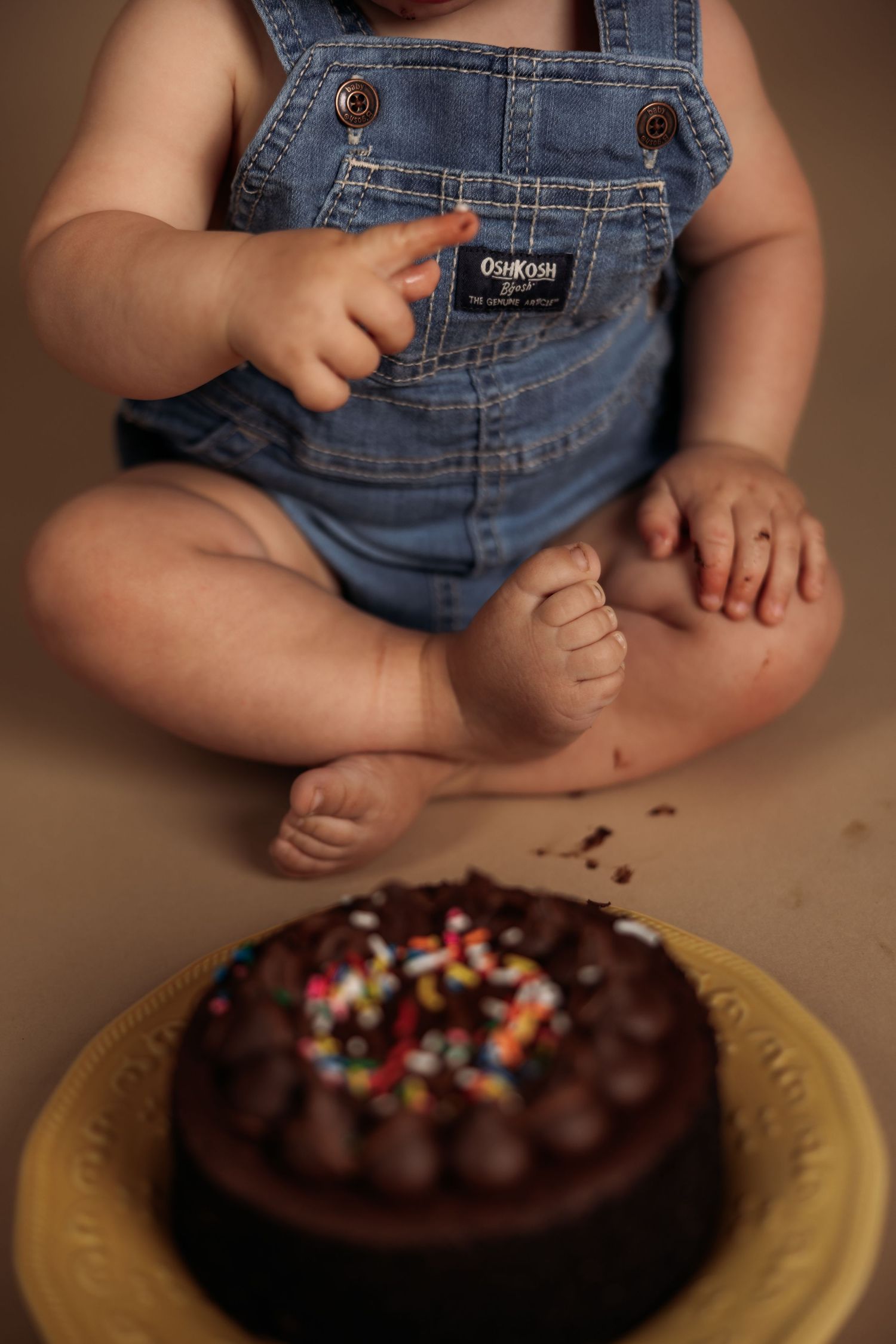 A baby in denim overalls sits next to a chocolate cake decorated with colorful sprinkles on a yellow plate.