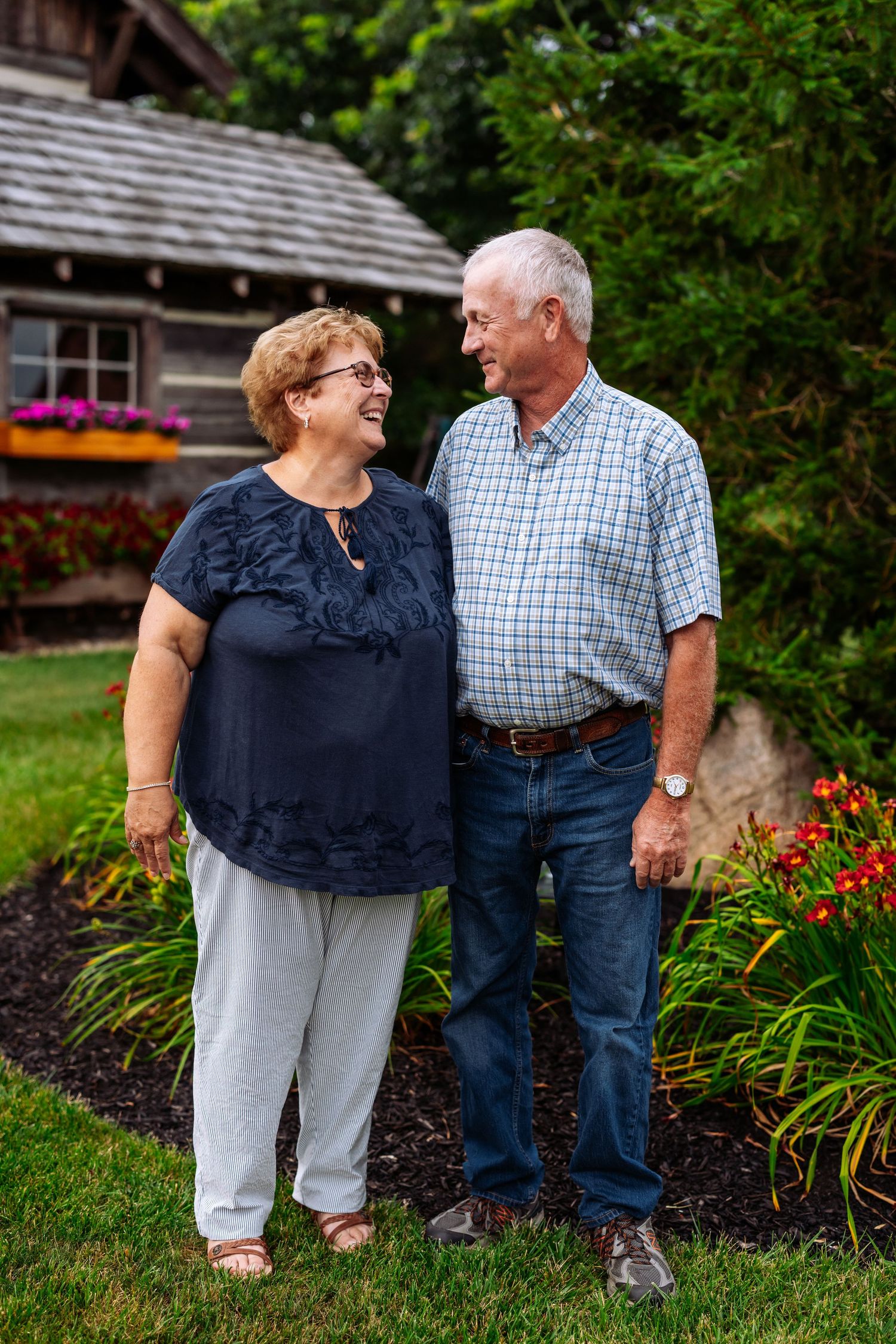 Senior couple smiling at each other while standing in garden with flowers and rustic cabin in background.