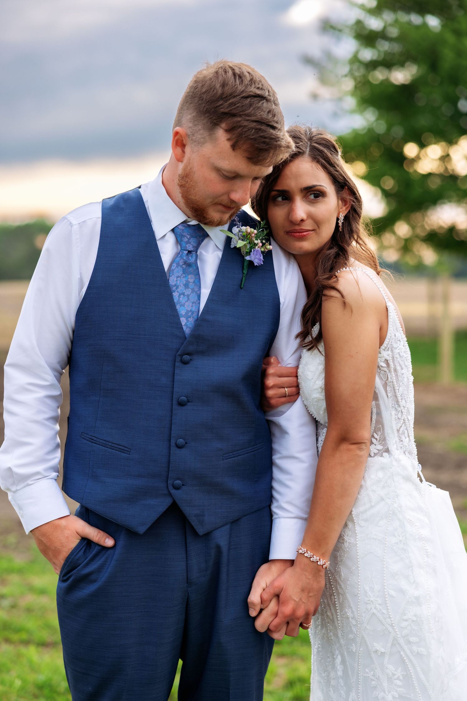 A bride in a white lace gown holds hands with a groom in a blue vest and tie, posing outdoors at sunset.