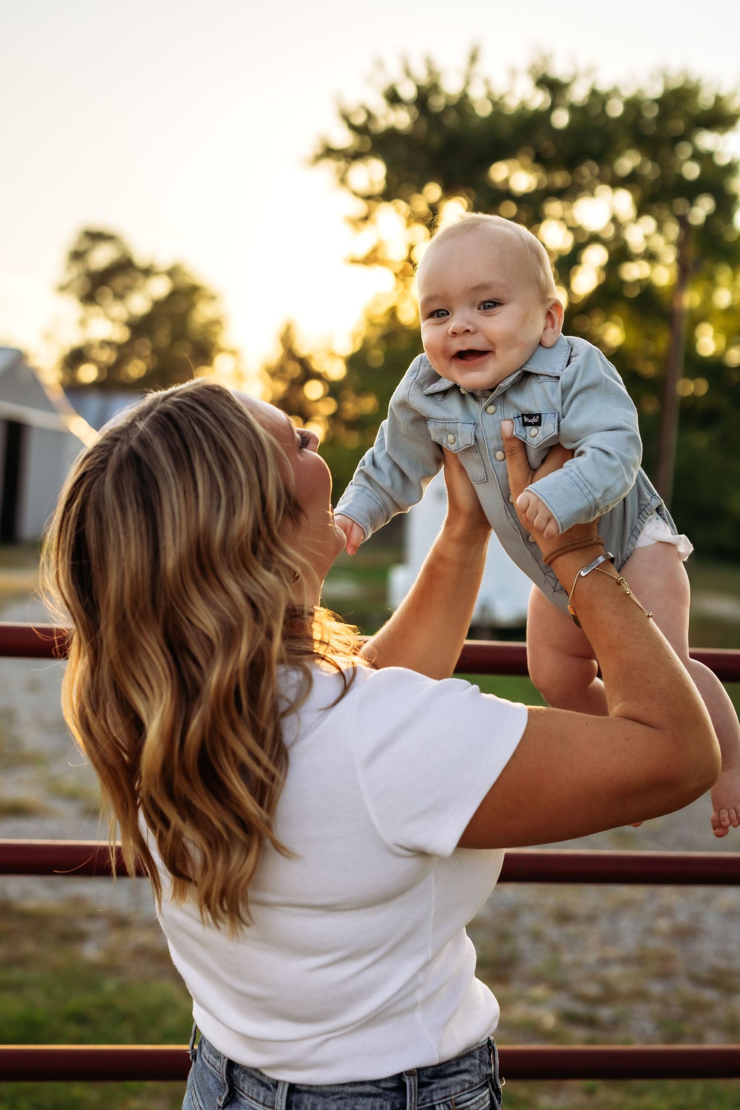Parent lifting joyful baby in denim outfit during sunset outdoors.