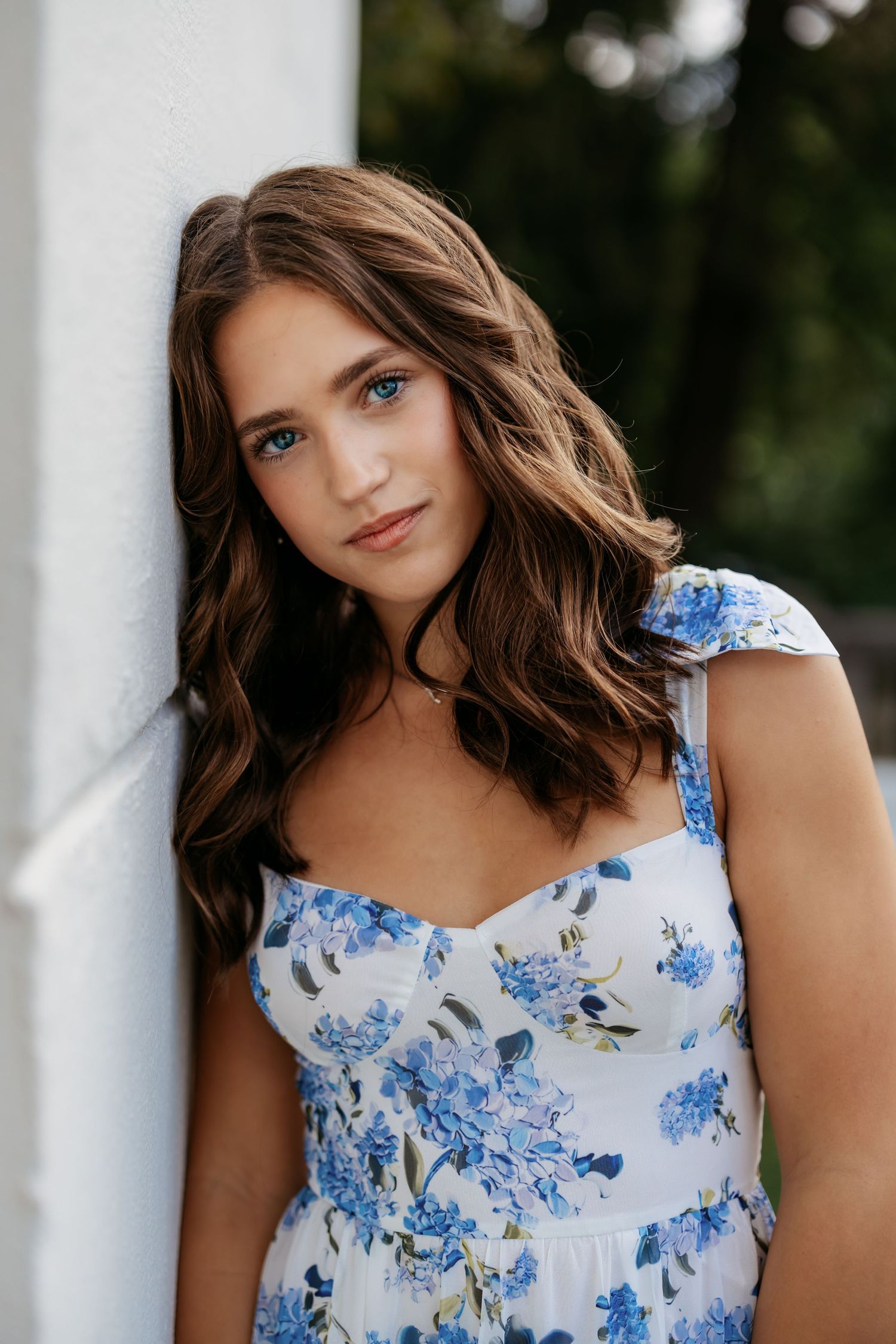 Woman in floral dress with wavy brown hair poses against white pillar outdoors.