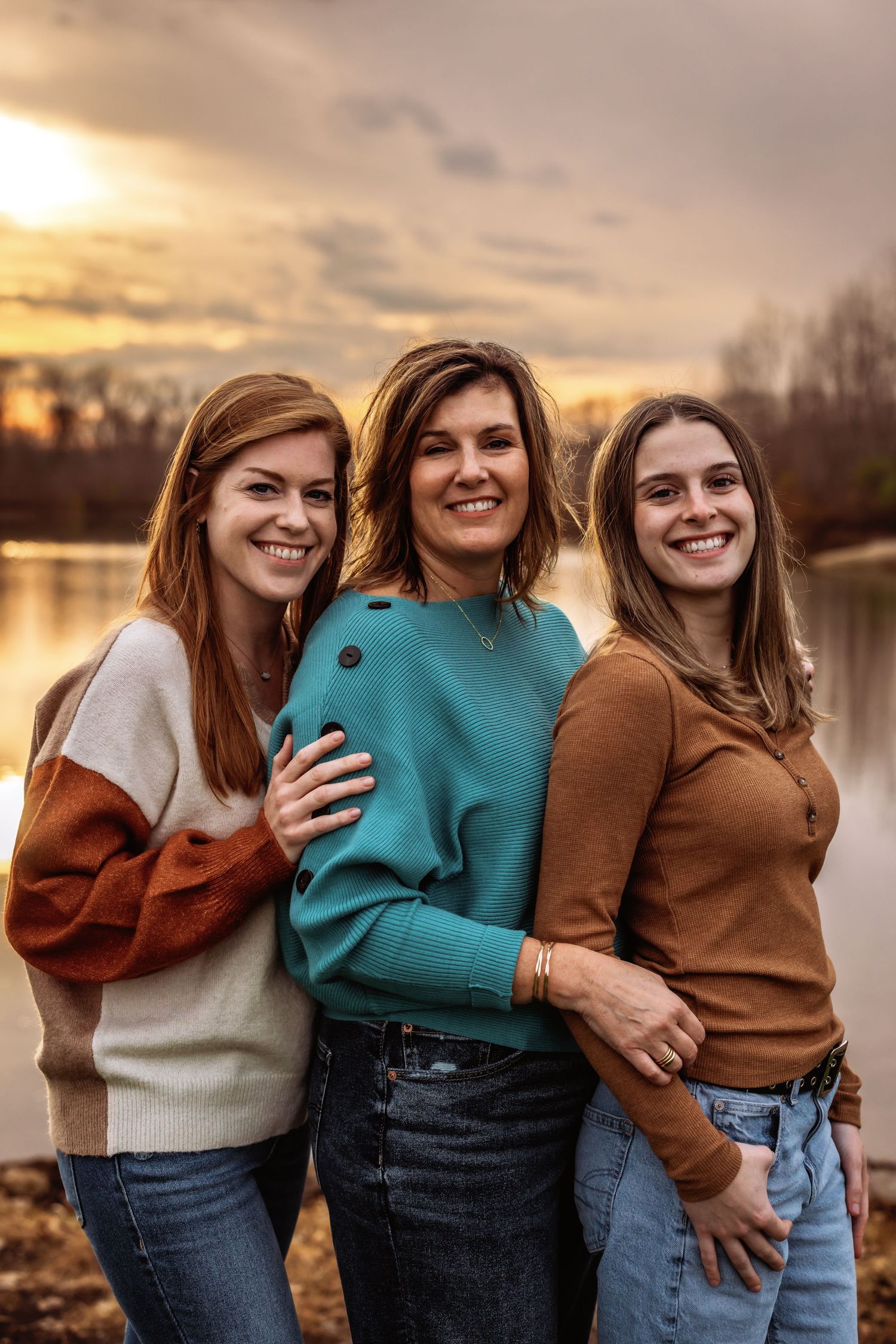 Three people in sweaters pose together by lake during golden sunset.