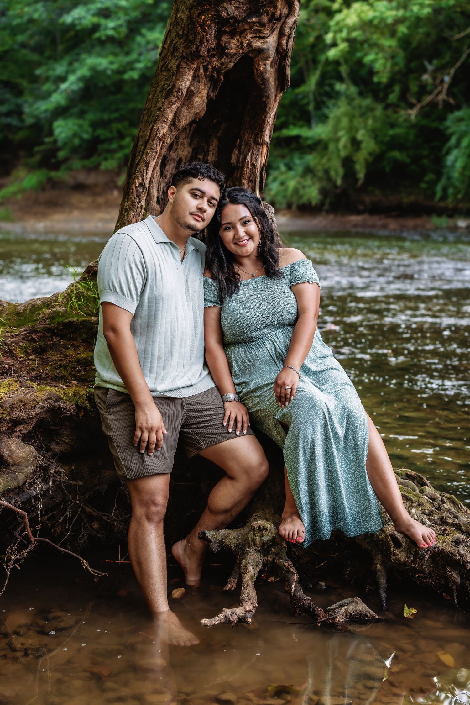 A couple sits together by a creek on exposed tree roots, sharing intimate moments in a natural forest setting.