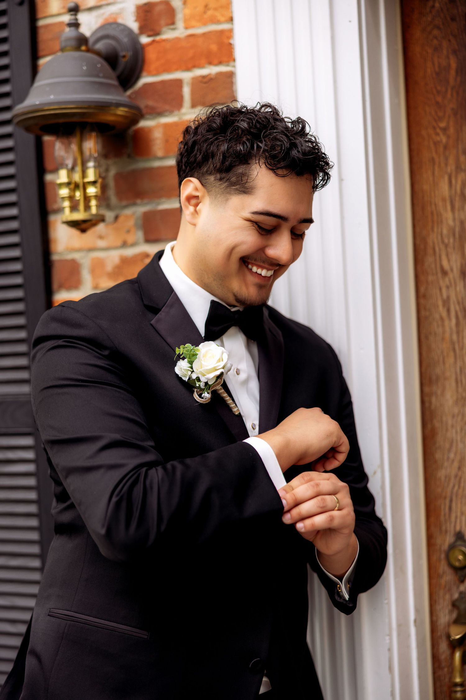 Smiling groom in black tuxedo with white boutonniere adjusting cufflinks outside brick building.