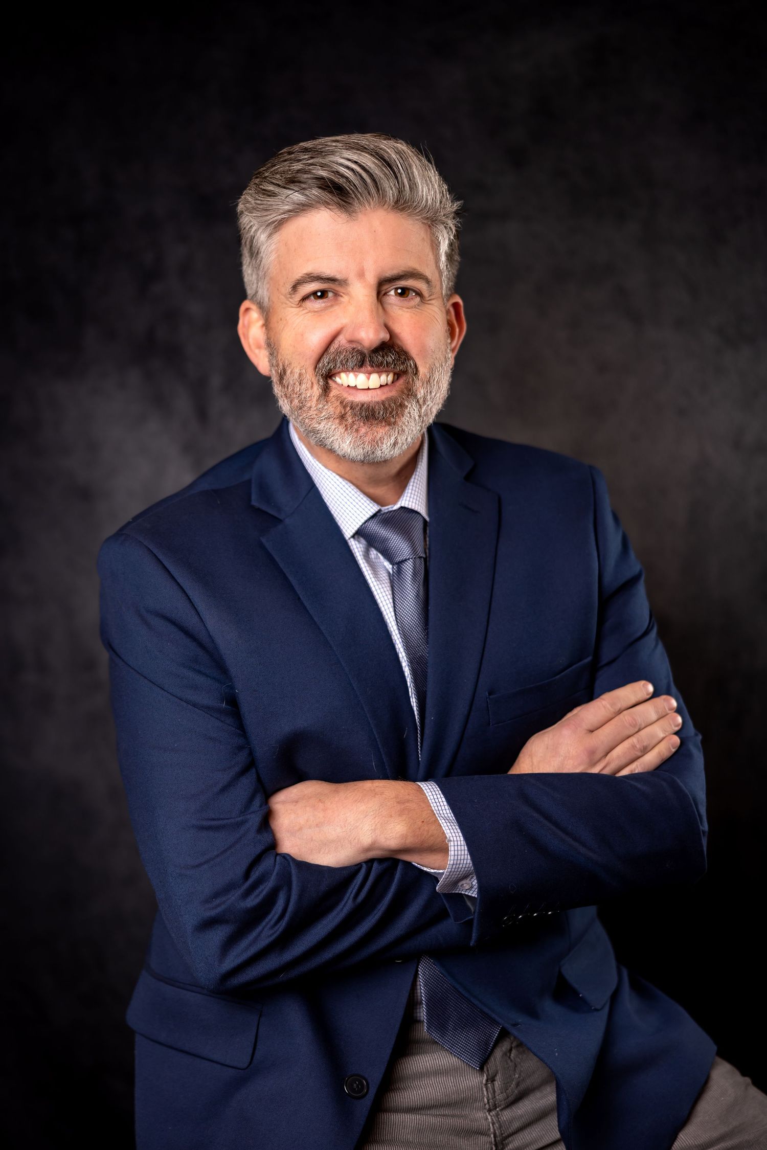 Professional headshot of person in navy suit and tie against dark background with confident expression.