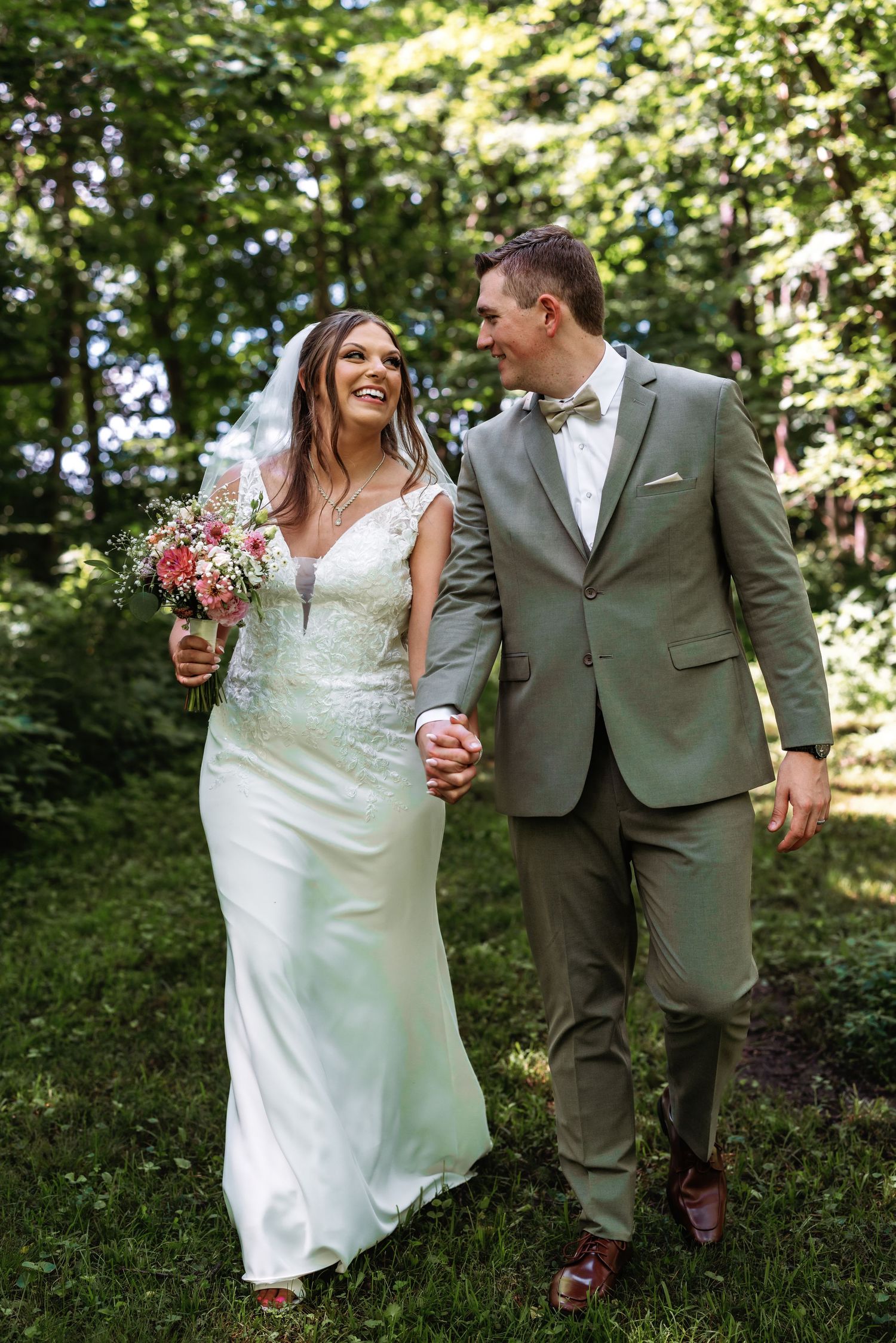 Newlywed couple walk hand in hand through wooded area, bride in white lace dress holding pink bouquet.