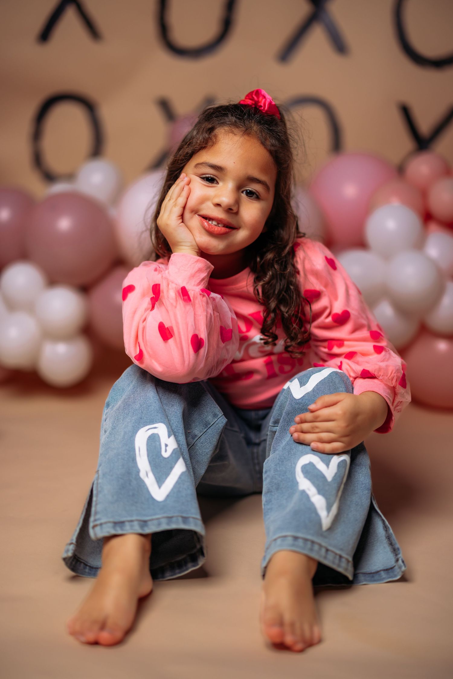 Child in pink sweater and heart-decorated jeans sitting against backdrop with pink balloons.