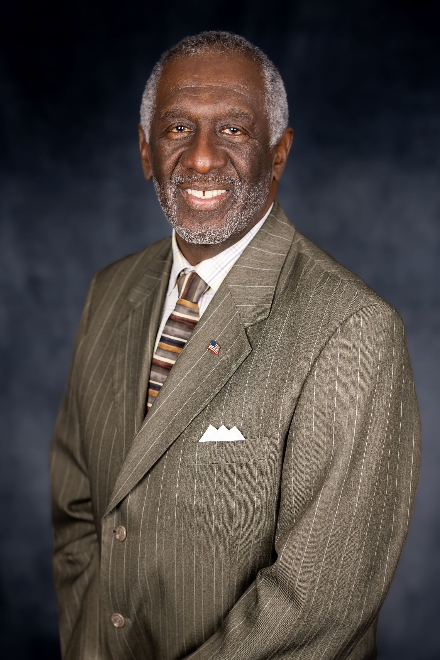 Professional portrait of a person wearing a pinstriped suit and striped tie against a dark background.