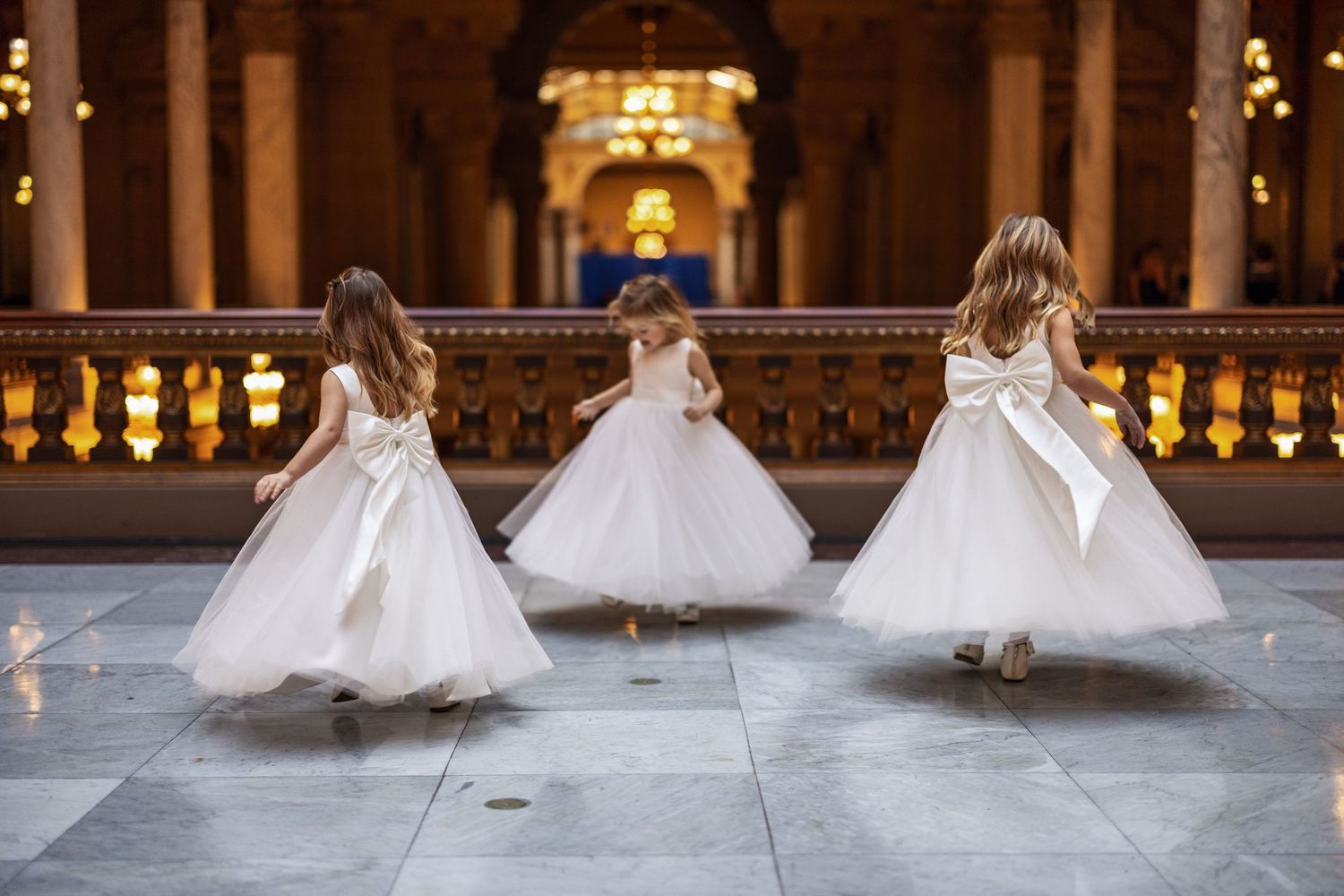 Three young girls in white dresses twirl and dance in an elegant hallway with ornate lighting and architecture.