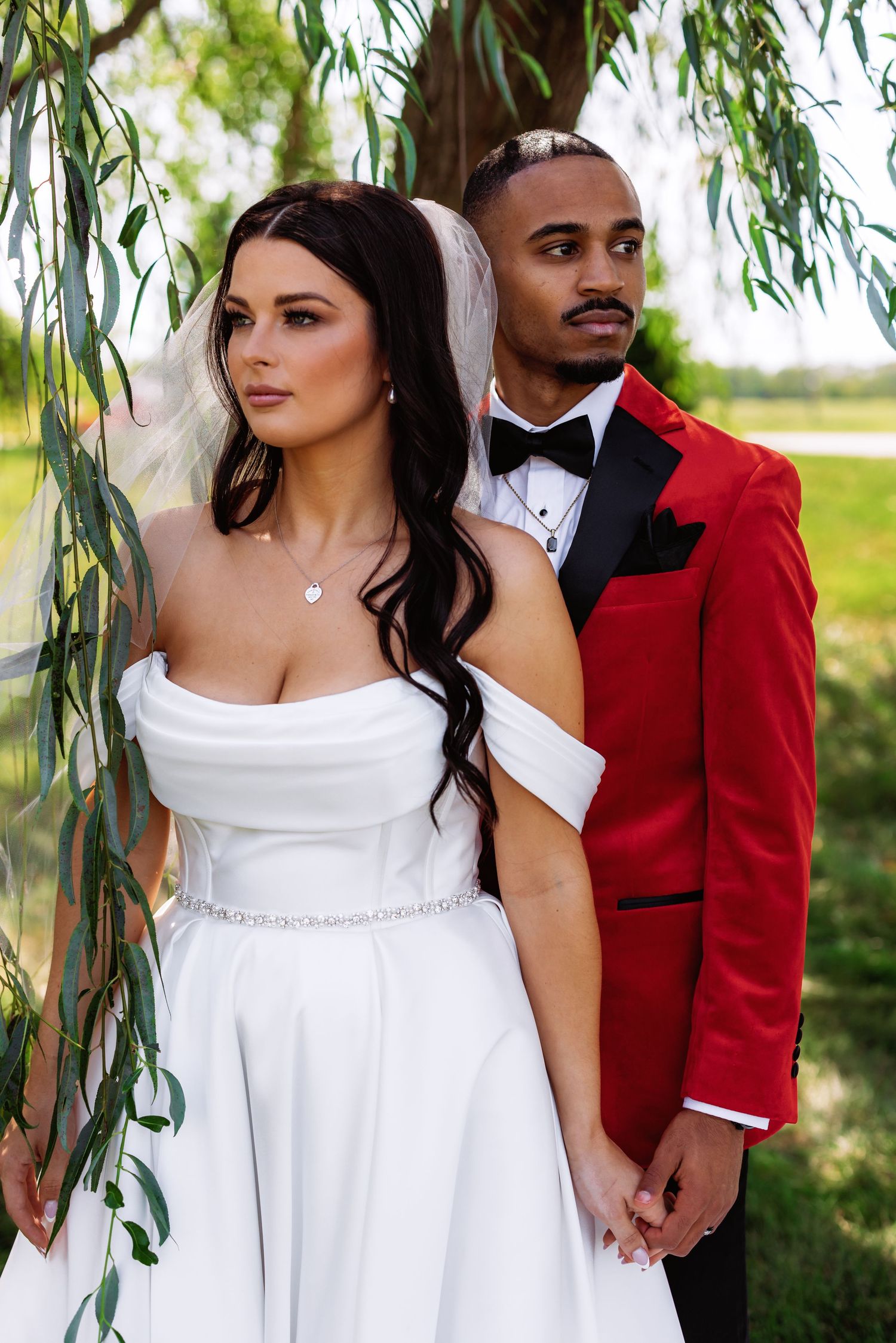 A stylish wedding photo under willow trees with a red tuxedo and off-shoulder white dress.