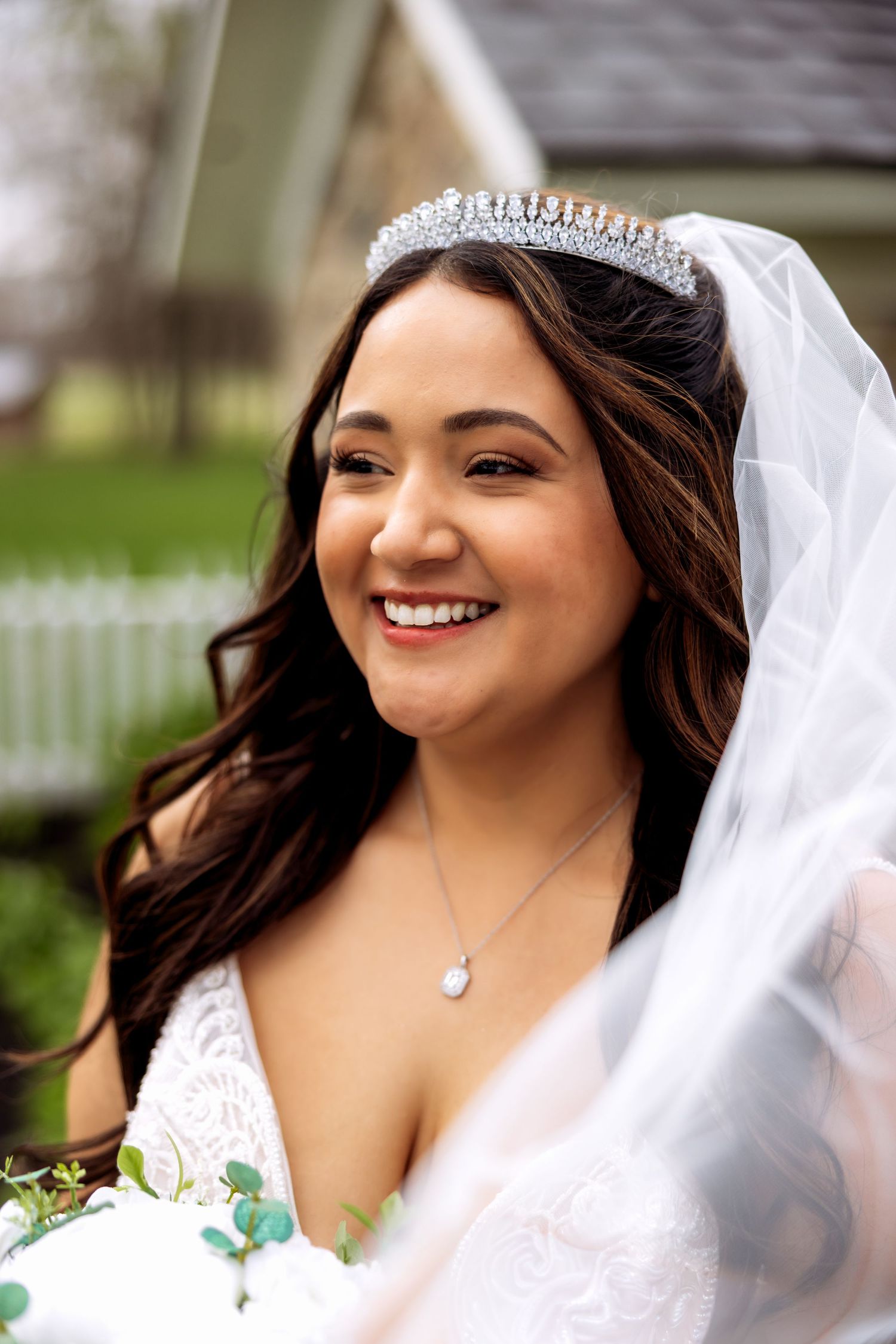 Smiling bride wearing a crystal tiara, veil, and white lace gown holds a bouquet outdoors.