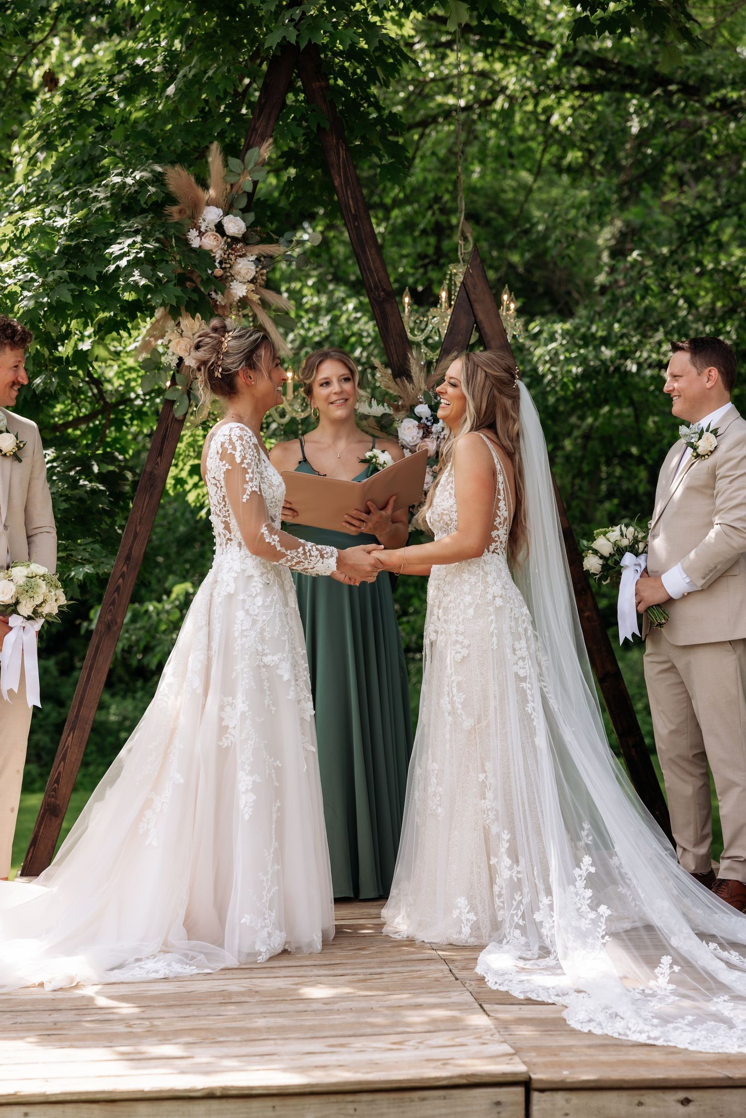 Two brides in white lace gowns participate in outdoor wedding ceremony with officiant and wedding party on wooden platform.
