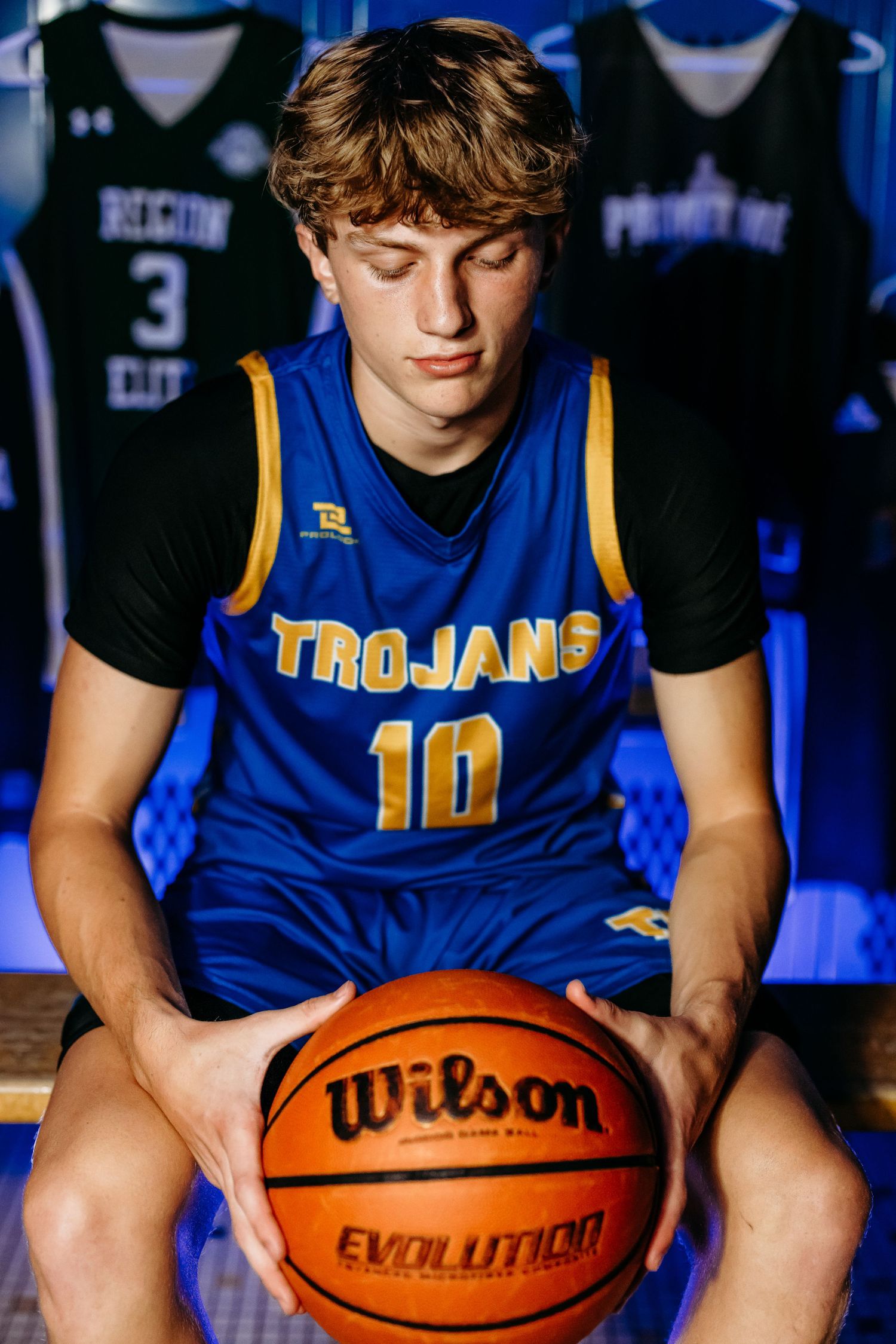High school athlete in blue Trojans jersey holding Wilson basketball in locker room setting.
