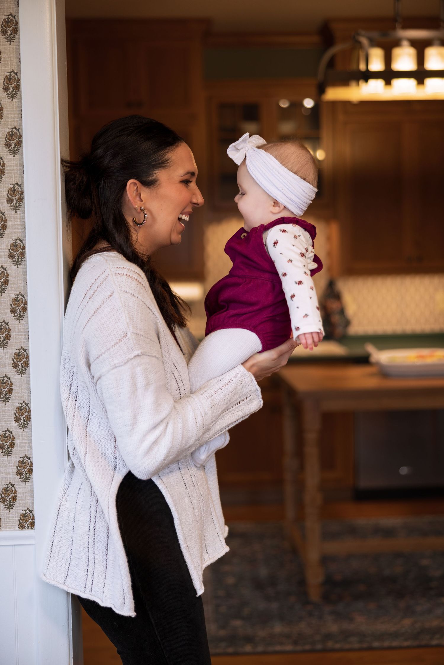 A tender moment shared between a woman in a white sweater and a baby wearing a burgundy outfit in a warmly lit home.