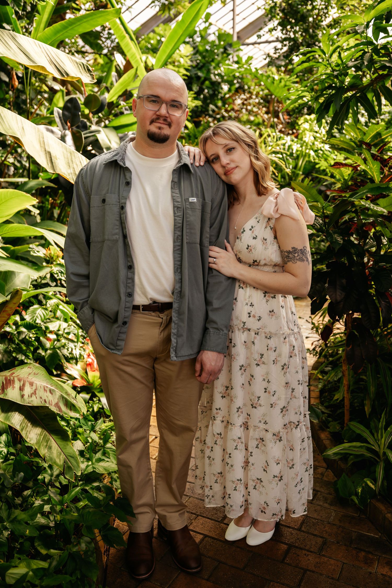 Couple posing in greenhouse surrounded by lush tropical plants and brick pathway.