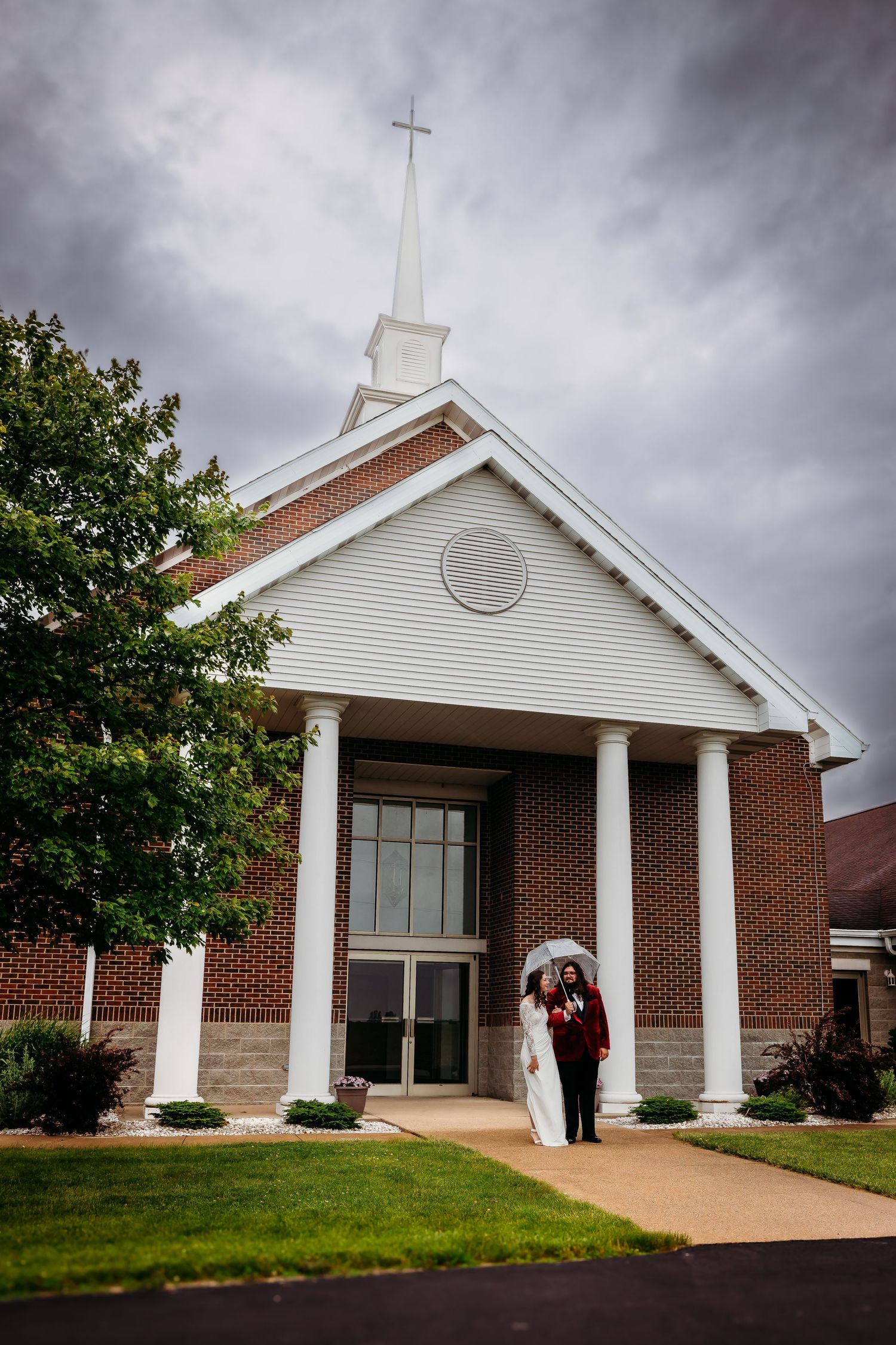 A wedding couple enters a white brick church with tall steeple under dramatic cloudy sky.