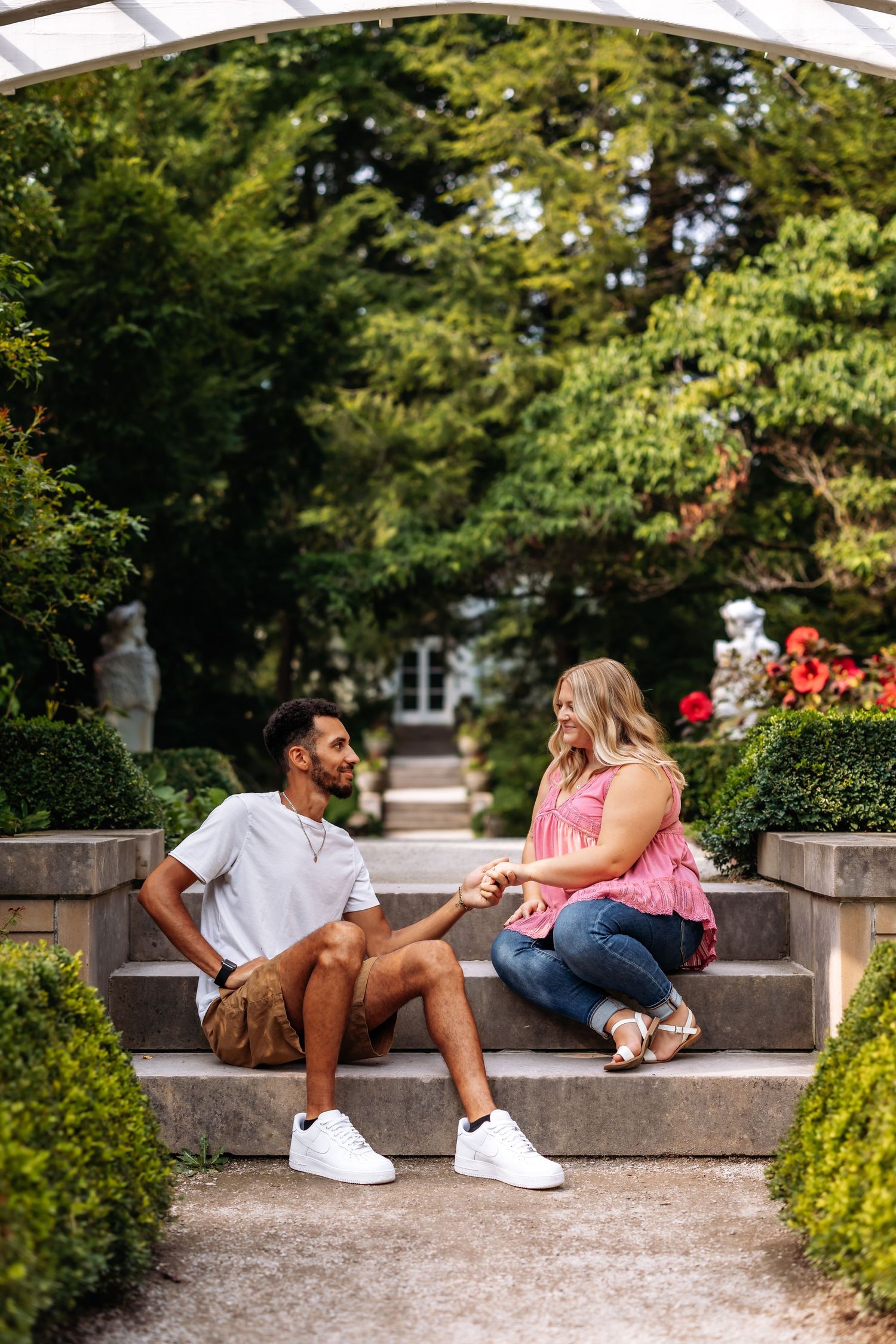 A couple sits on stone steps under an archway surrounded by manicured garden bushes.