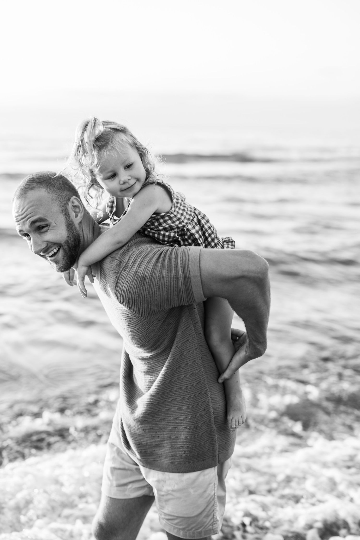 Playful beach moment with a piggyback ride along the ocean shore in black and white.