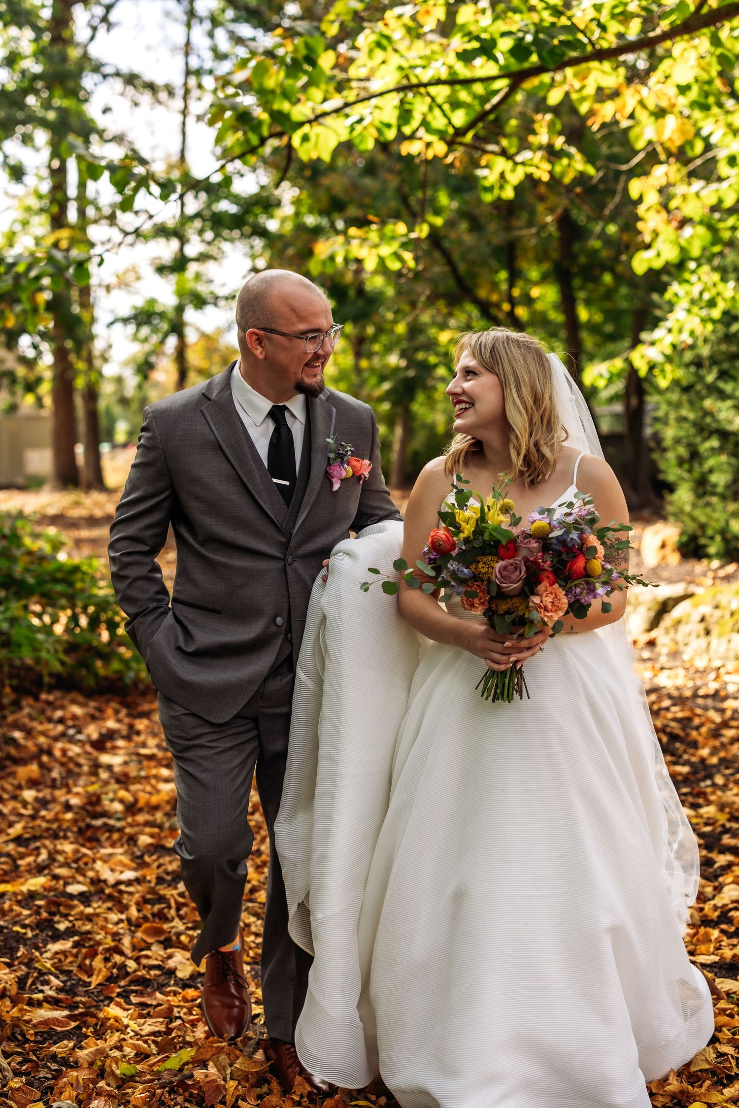 A wedding couple walks through autumn leaves in a wooded setting with colorful florals.
